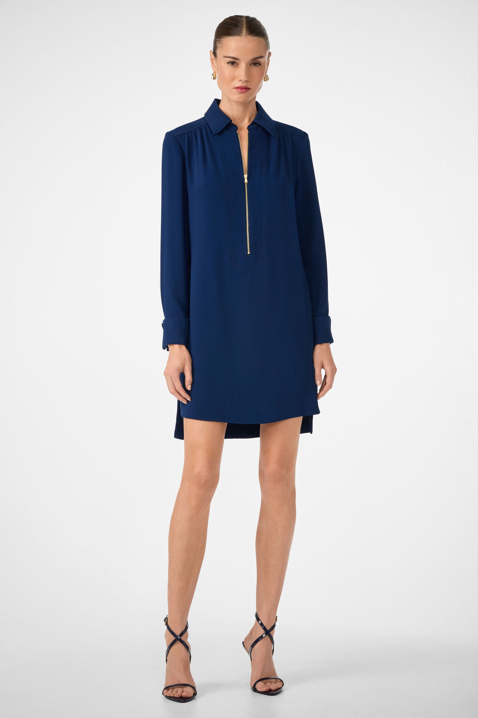 A woman stands against a plain background wearing the Darya Long-Sleeve Dress in Ink, featuring a gold zipper at the neckline and black high-heeled sandals.