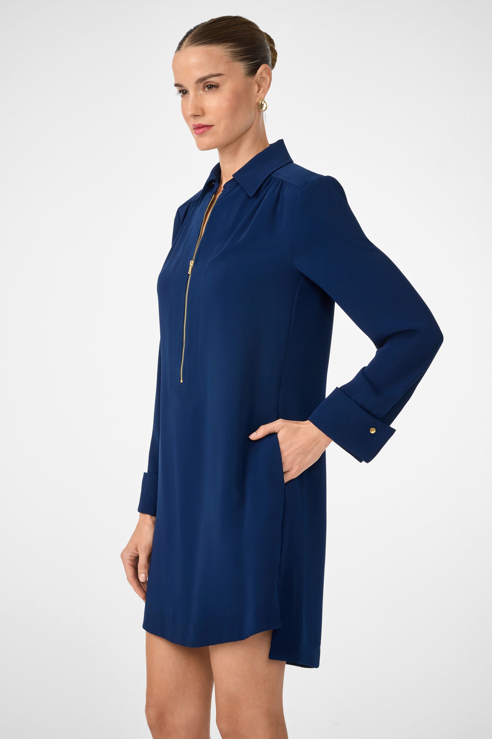 A woman with brown hair in a sleek bun wears the Darya Long-Sleeve Dress, a navy blue shift style featuring a gold zipper and pockets, posing sideways with one hand in her pocket against a plain background.