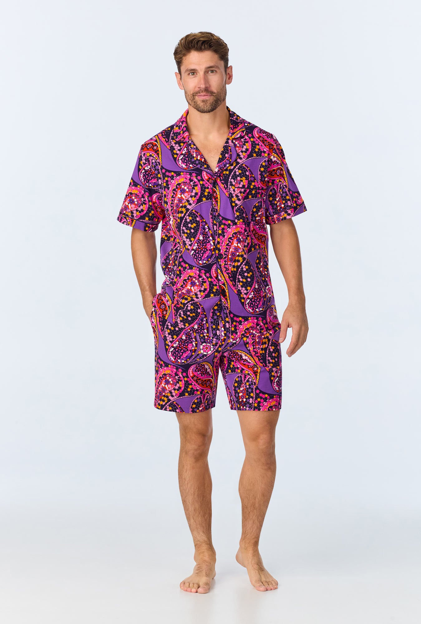 A man stands barefoot against a plain background, wearing the Tangled Paisley Mens Short Sleeve Boxer Short Jersey PJ Set in multi, which features a vibrant paisley print with a matching button-up shirt and shorts.