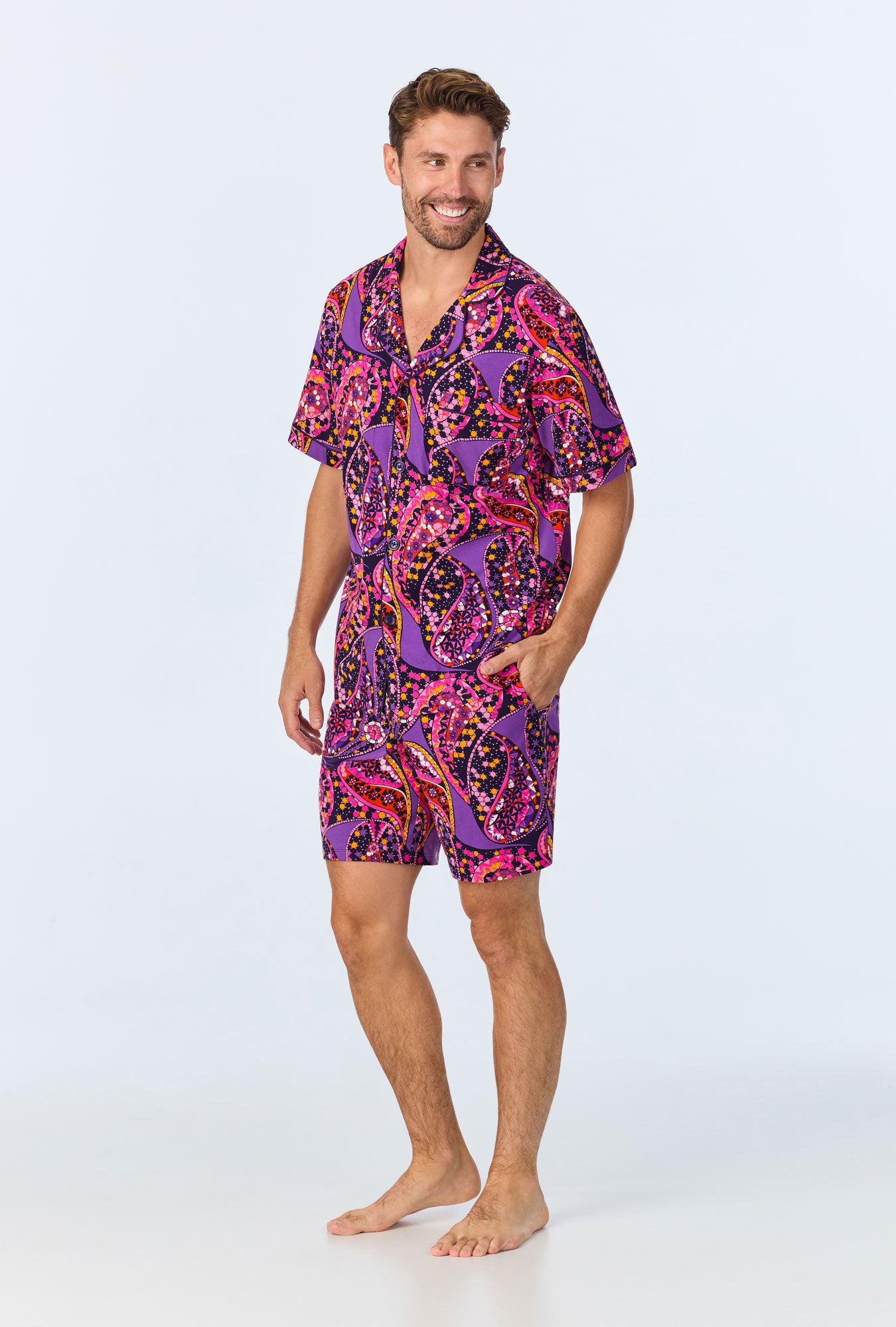 A man smiles barefoot in the Tangled Paisley Mens Short Sleeve Boxer Short Jersey PJ Set, featuring a bold purple, pink, orange, and black paisley print on a light background—perfect comfy sleepwear in organic cotton.