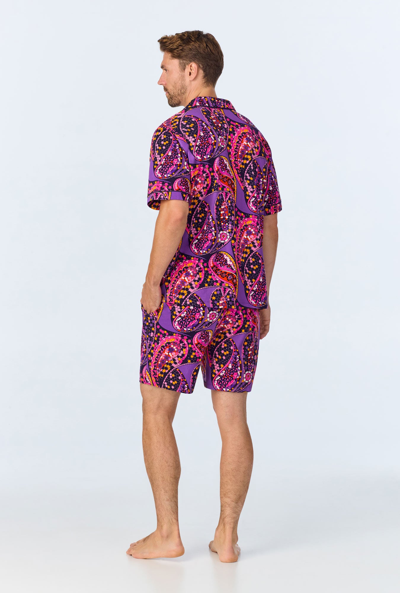 A man stands barefoot facing away, wearing the Tangled Paisley Mens Short Sleeve Boxer Short Jersey PJ Set with a vibrant purple and pink pattern, set against a plain light background.