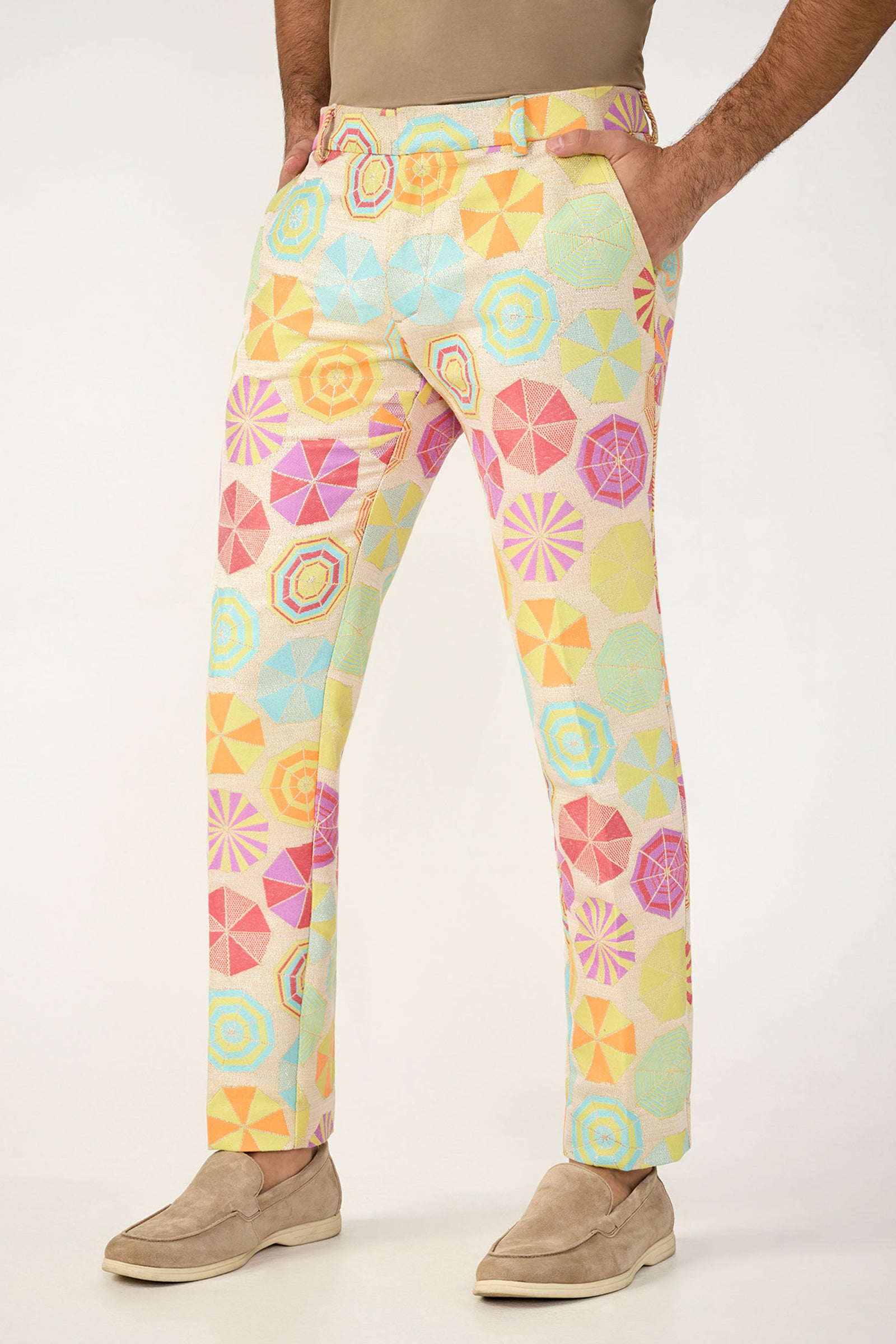 A person wearing Clyde Slim Trouser - Parapluie Jacquard, featuring a pastel geometric pattern, with beige shoes and a light brown shirt. Their hands are in their pockets.