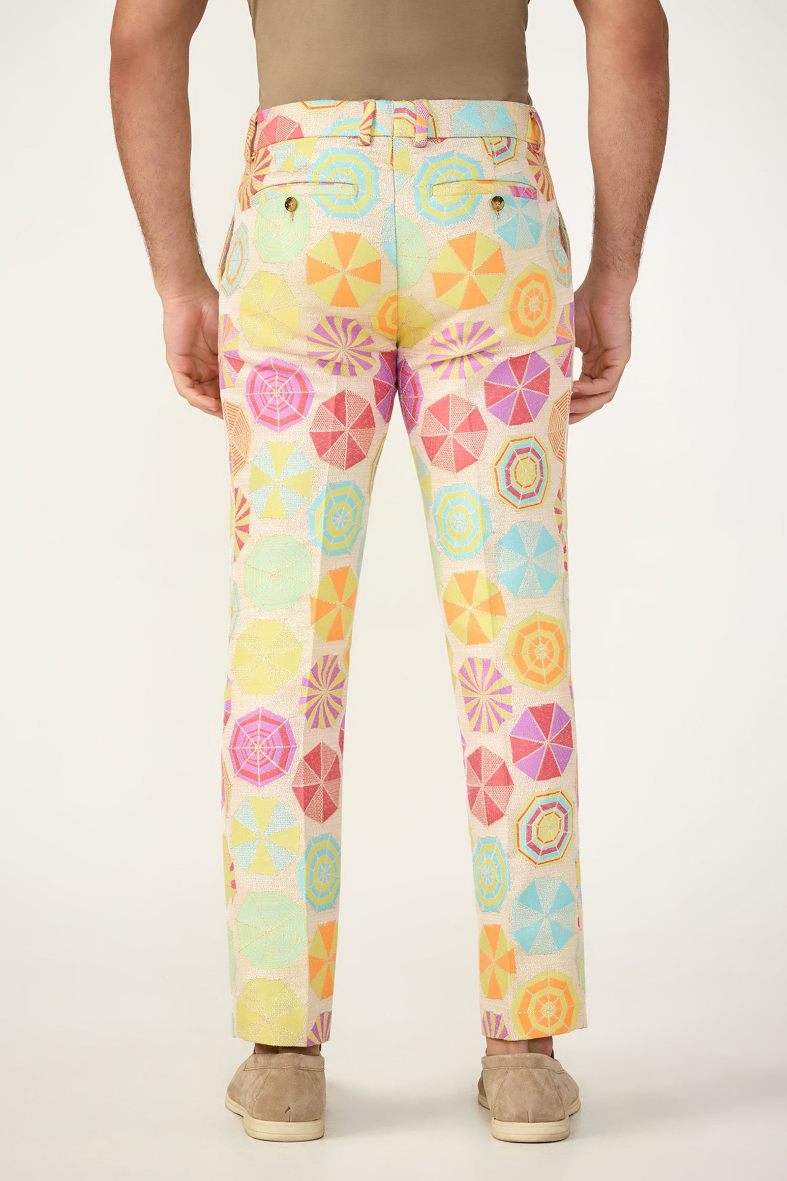 A person faces away, wearing the Clyde Slim Trouser in Parapluie Jacquard—a vibrant pastel geometric and umbrella pattern—paired with tan shoes and a beige shirt. The background is plain white.