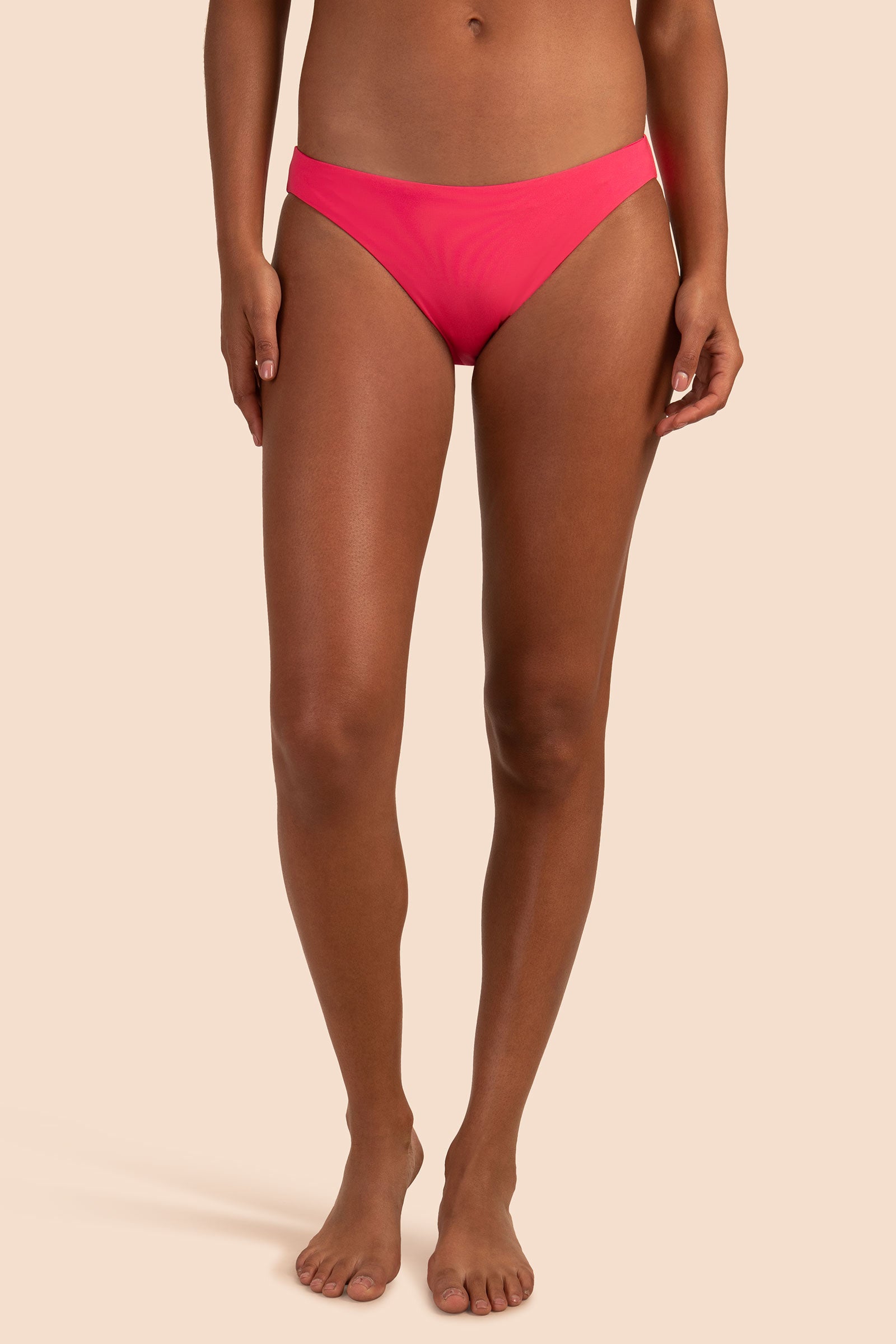 A woman stands barefoot against a beige background, modeling the Women's Monaco Low Rise French Cut Swim Bottom in bright pink. Her upper body and face are not visible, with arms relaxed at her sides.