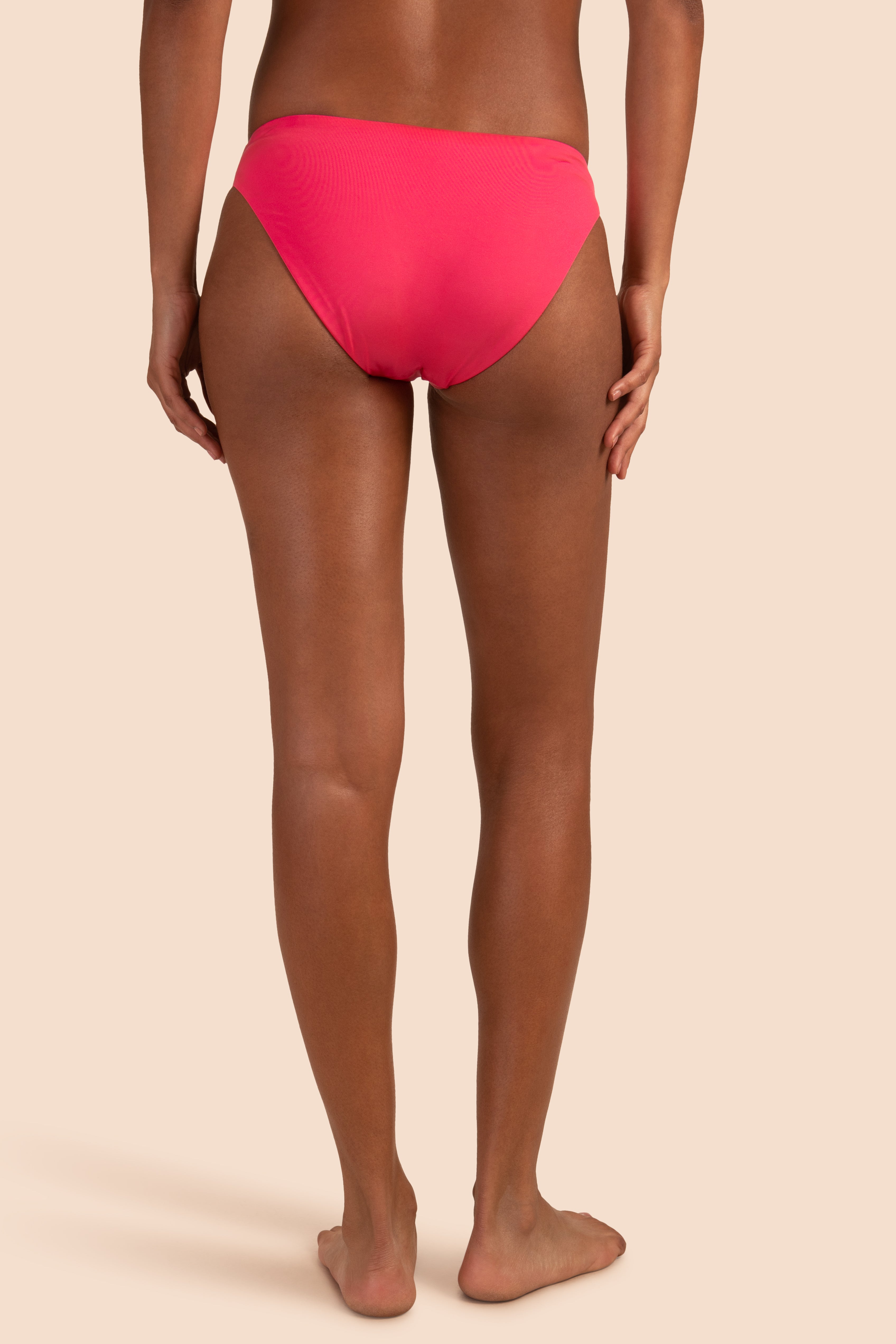 A person stands barefoot on a neutral background, shown from the back, wearing Women's Monaco Low Rise French Cut Swim Bottom in bright pink.