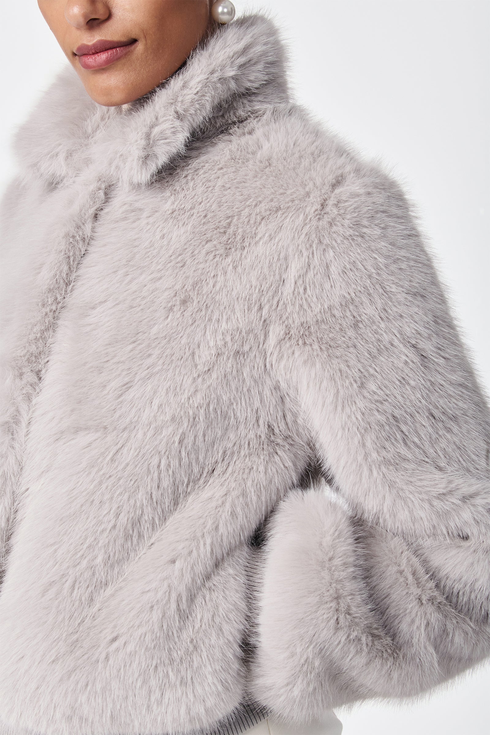A woman models the Morris Short Faux Fur Coat in light gray, featuring a high collar and soft texture, with one hand in her pocket and a visible pearl earring against a plain white background.
