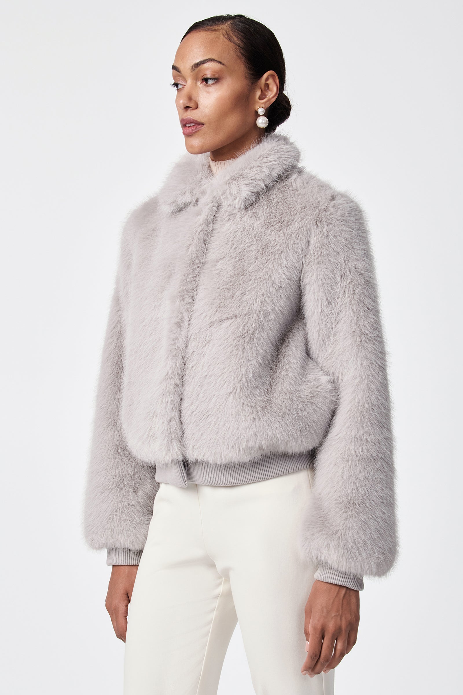 A person wearing the Morris Short Faux Fur Coat in light gray with a collar and ribbed cuffs, paired with white pants, stands against a plain white background.