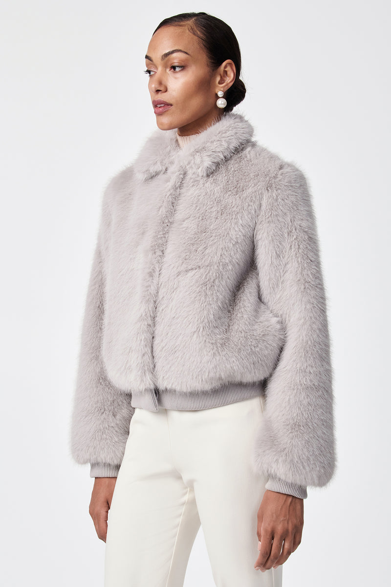 A person wearing the Morris Short Faux Fur Coat in light gray with a collar and ribbed cuffs, paired with white pants, stands against a plain white background.