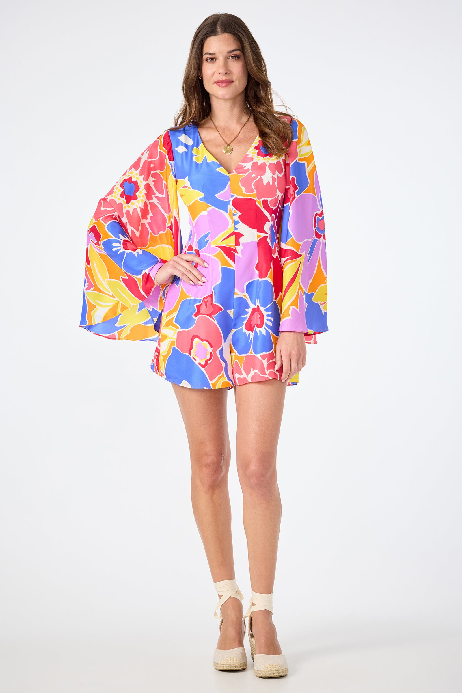A woman wears the Miera V-Neck Romper in Soie Paradis, featuring a colorful floral print and wide sleeves, paired with white platform espadrille sandals against a plain white background.
