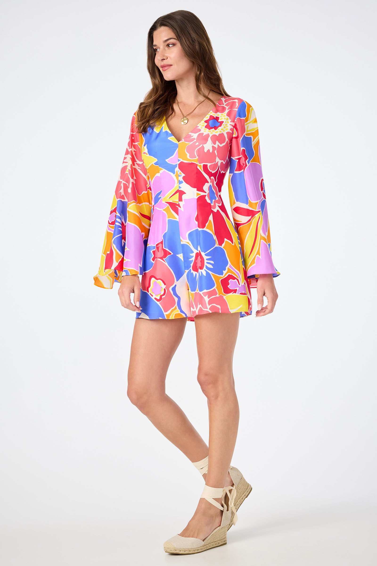 A woman stands confidently in the Miera V-Neck Romper - Soie Paradis, featuring long flared sleeves and a vibrant floral print. She completes the look with beige wedge espadrilles against a plain white background.