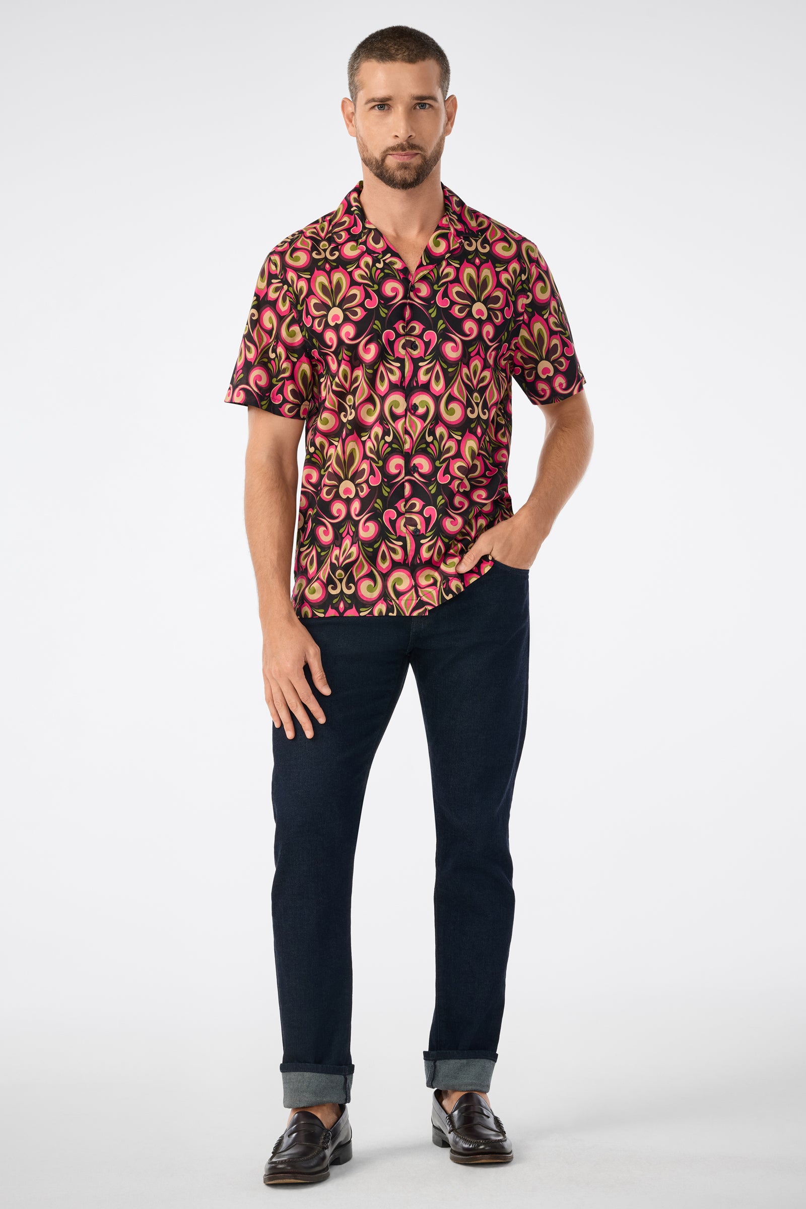 A man stands against a plain background wearing the Felix Camp Shirt - Sanctuary, a short-sleeve multicolored patterned shirt, paired with dark blue cuffed pants and black dress shoes. One hand is in his pocket as he looks straight ahead.