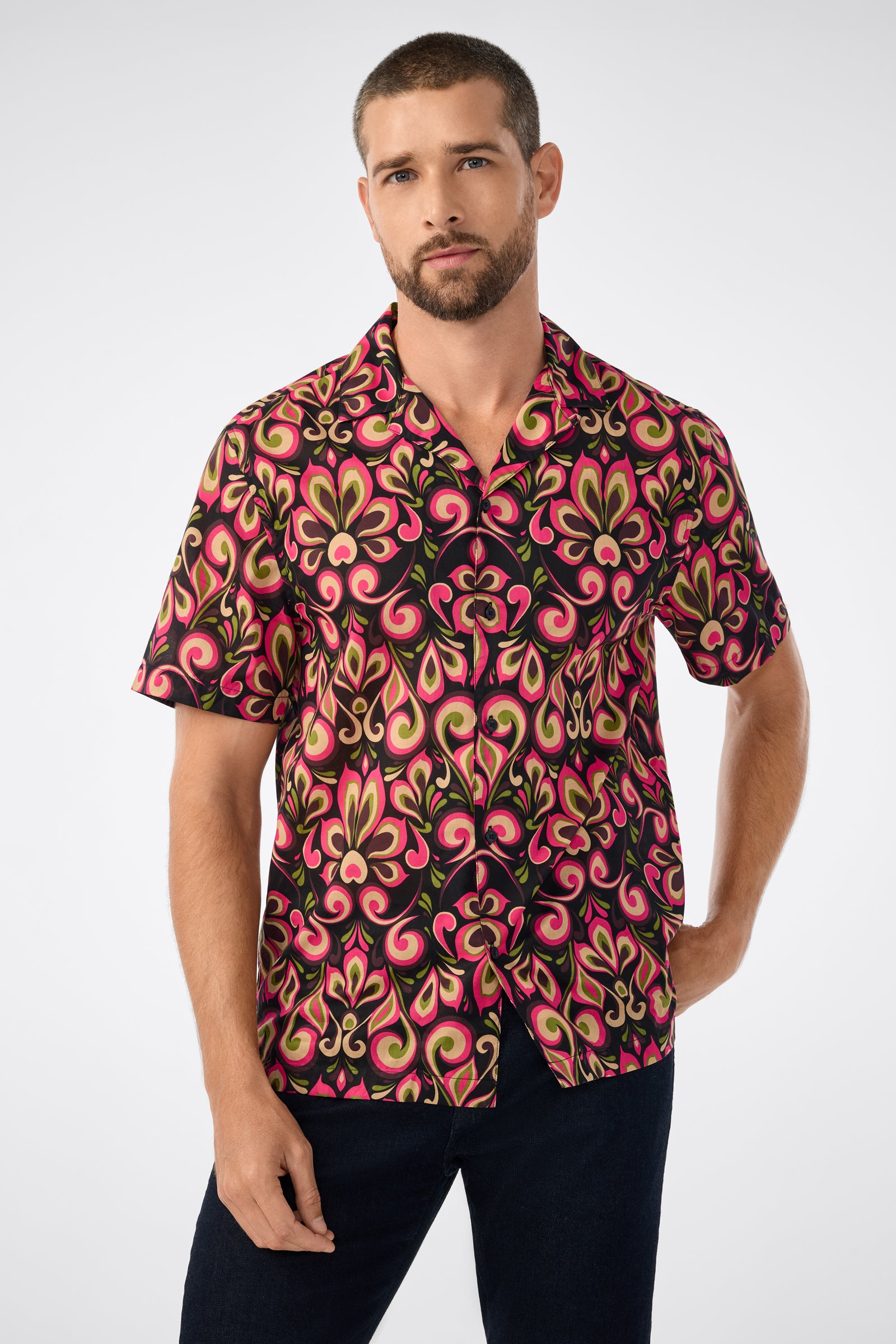 A man with short hair and a trimmed beard wears the Felix Camp Shirt in Sanctuary, featuring a bold, colorful retro pattern. He stands with one hand in his pocket against a plain background.