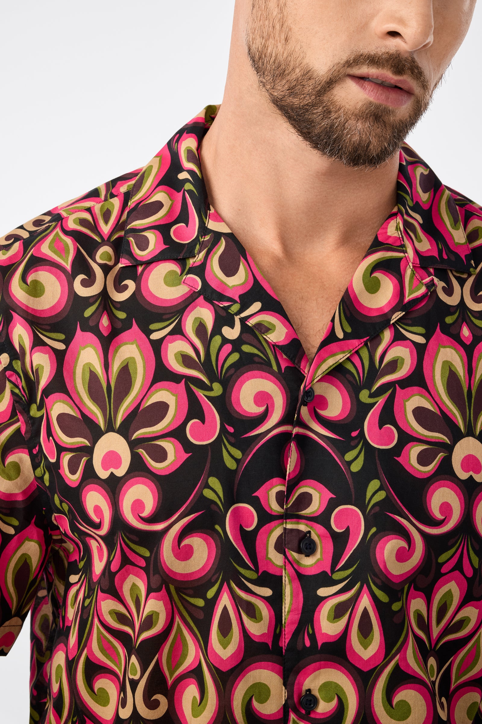 A man with a trimmed beard wears the Felix Camp Shirt - Sanctuary, featuring vibrant pink, green, and beige swirls and floral patterns. The image partially shows his face, highlighting the shirt’s bold design.