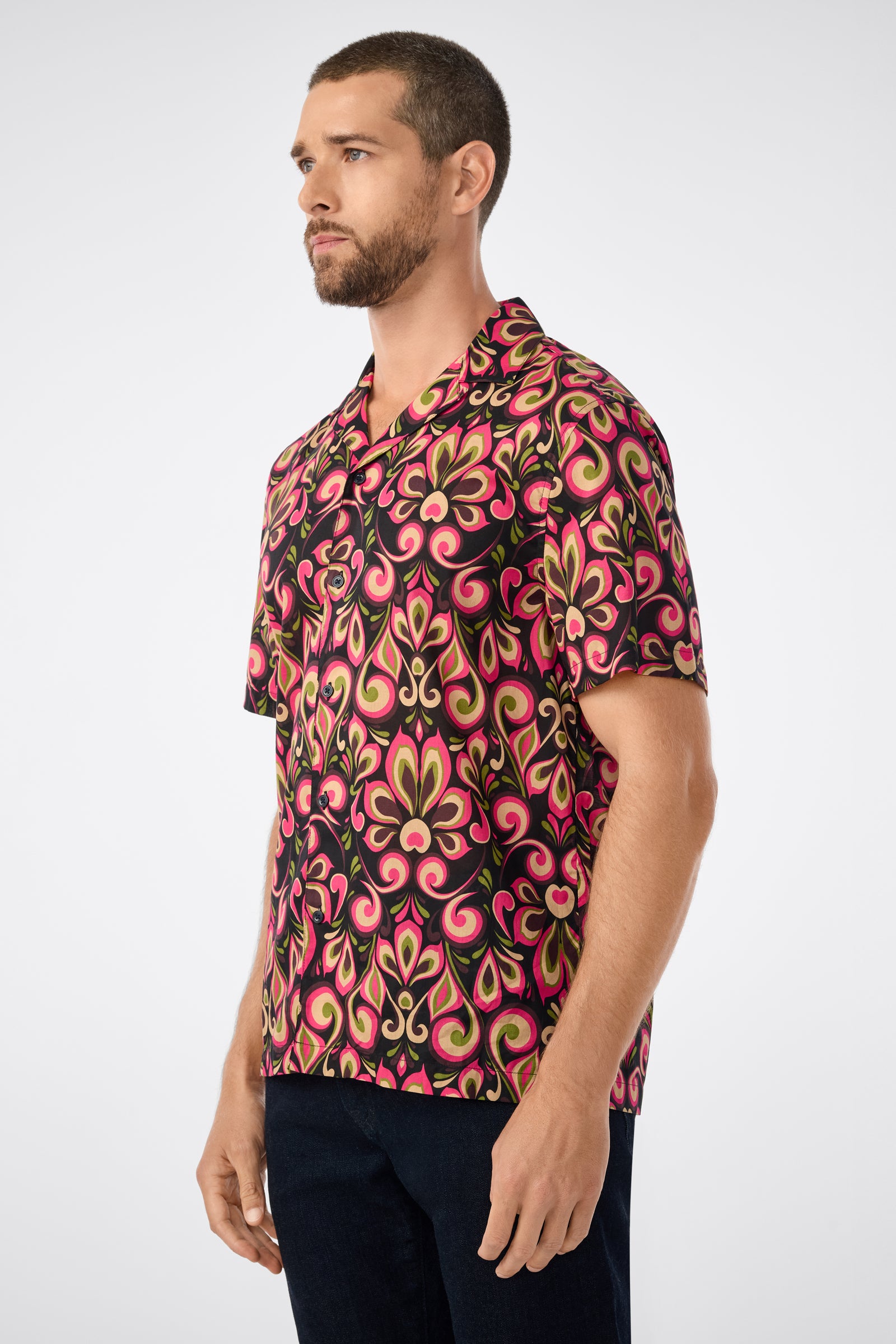 A man with short hair and a beard wears the Felix Camp Shirt - Sanctuary, a short-sleeved shirt with pink, green, and yellow patterns on a black background. He faces left against a plain light backdrop.