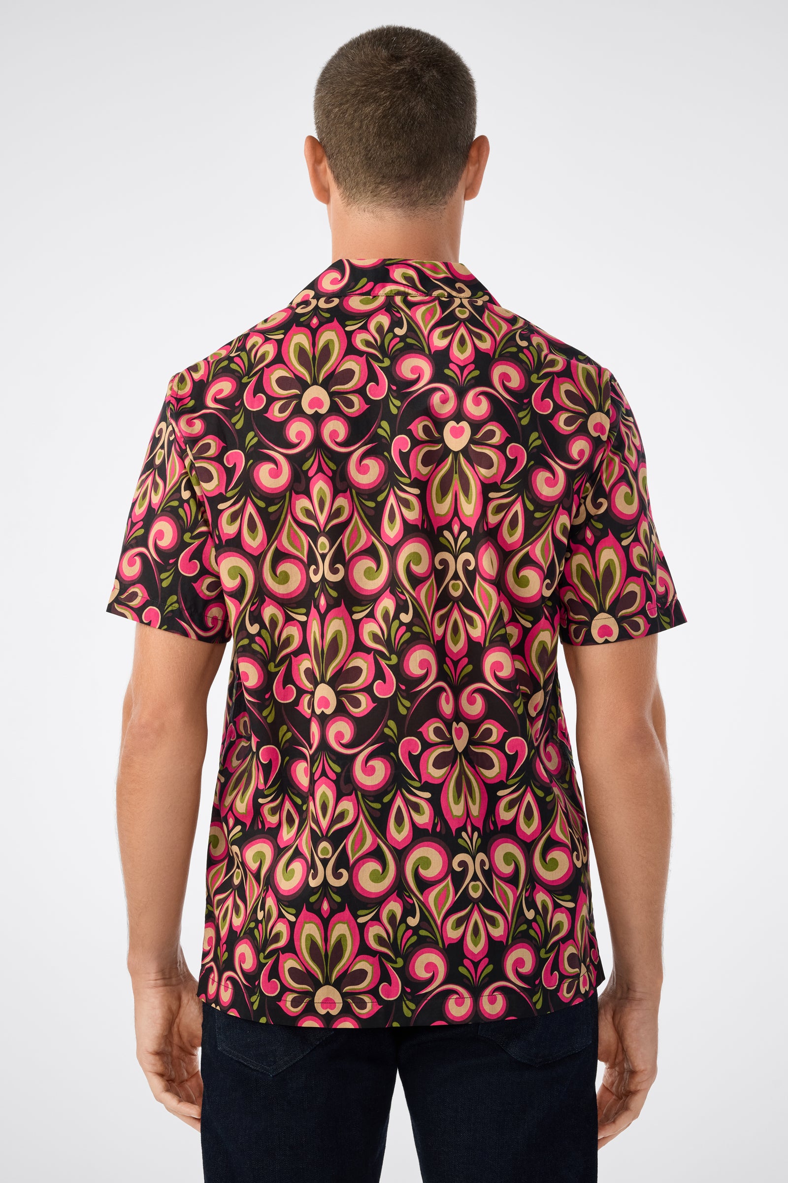 A person with short hair is shown from behind wearing the Felix Camp Shirt - Sanctuary, which features a bold swirling pink, green, and black pattern, along with dark pants.