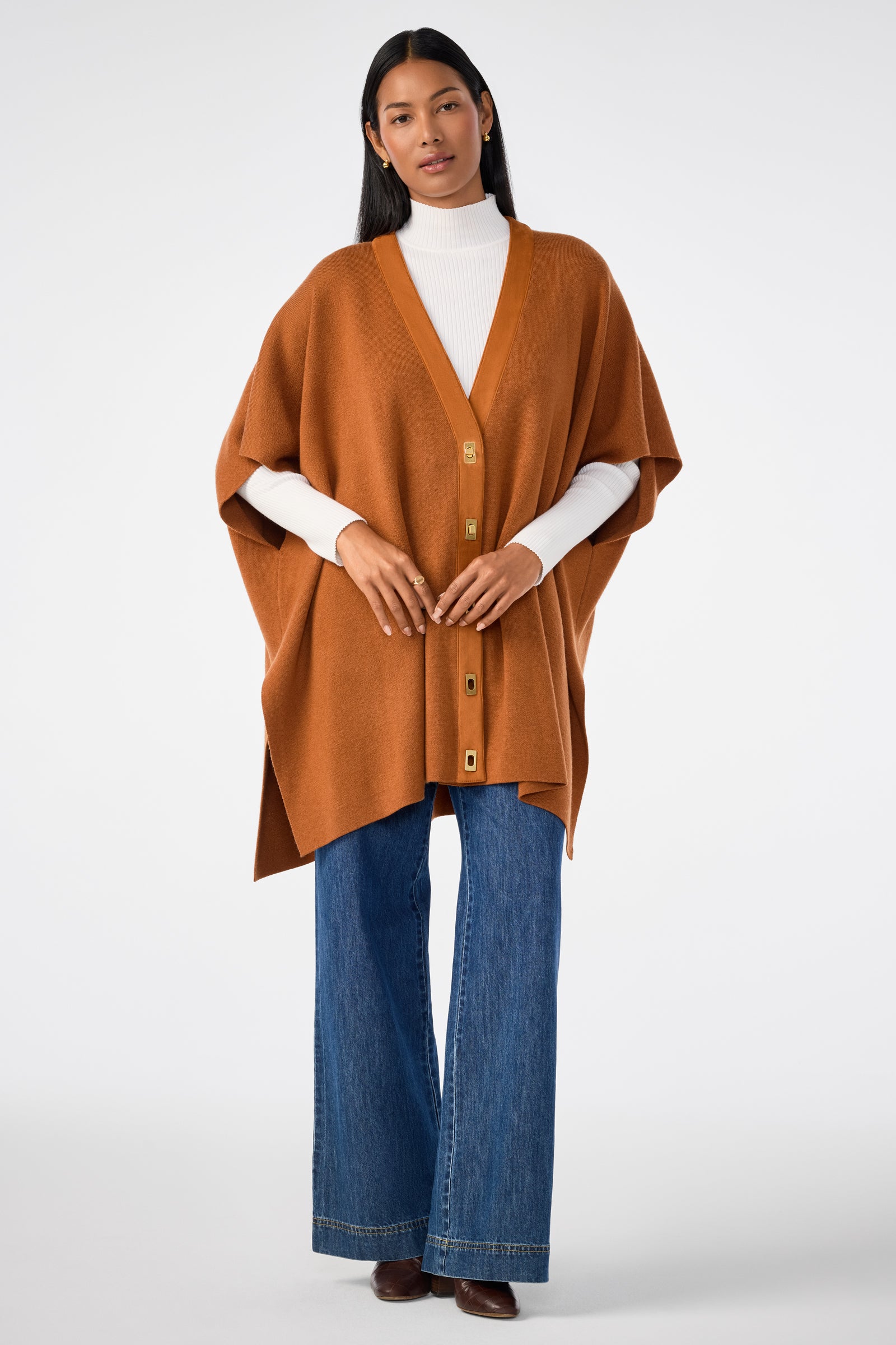 A woman with long dark hair and hoop earrings stands against a plain background, wearing the Sapphire Poncho over a white turtleneck, paired with blue wide-leg jeans and brown shoes.