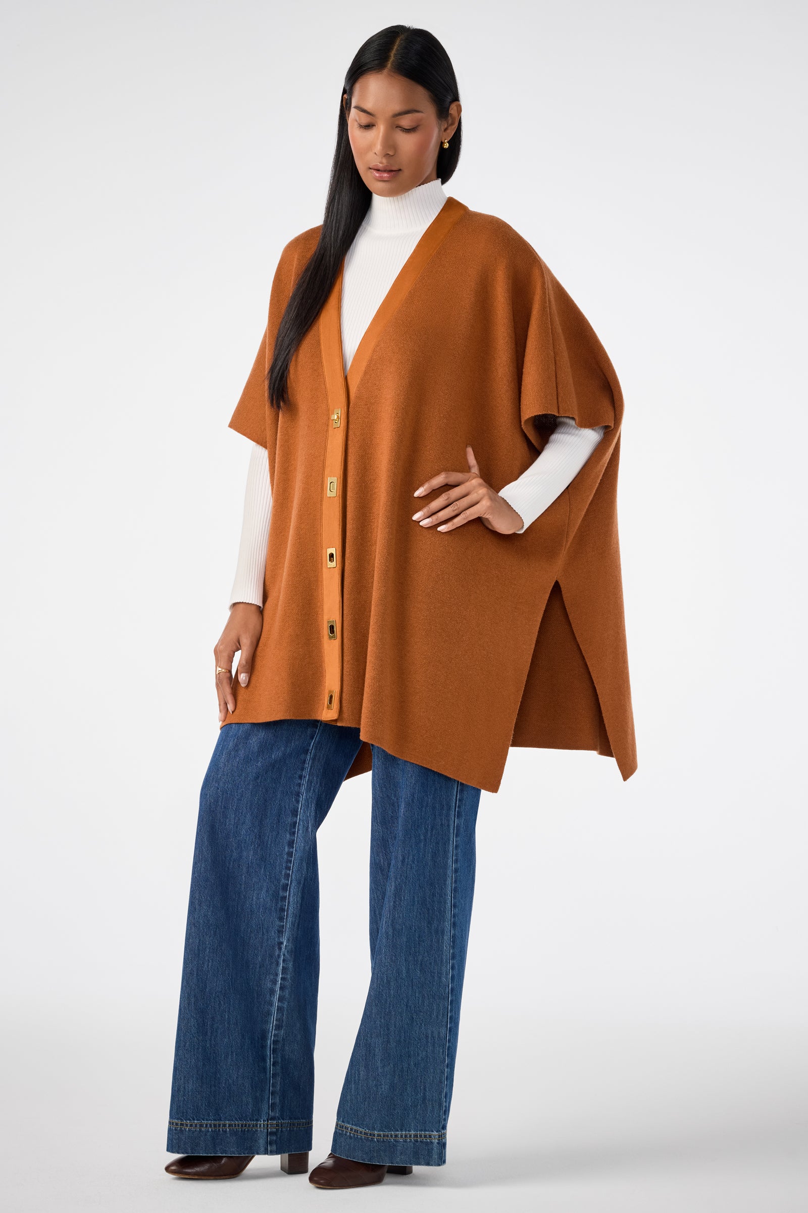 A woman poses against a white background wearing the Sapphire Poncho, a rust-colored button-up piece over a white turtleneck, paired with wide-leg blue jeans and brown shoes.