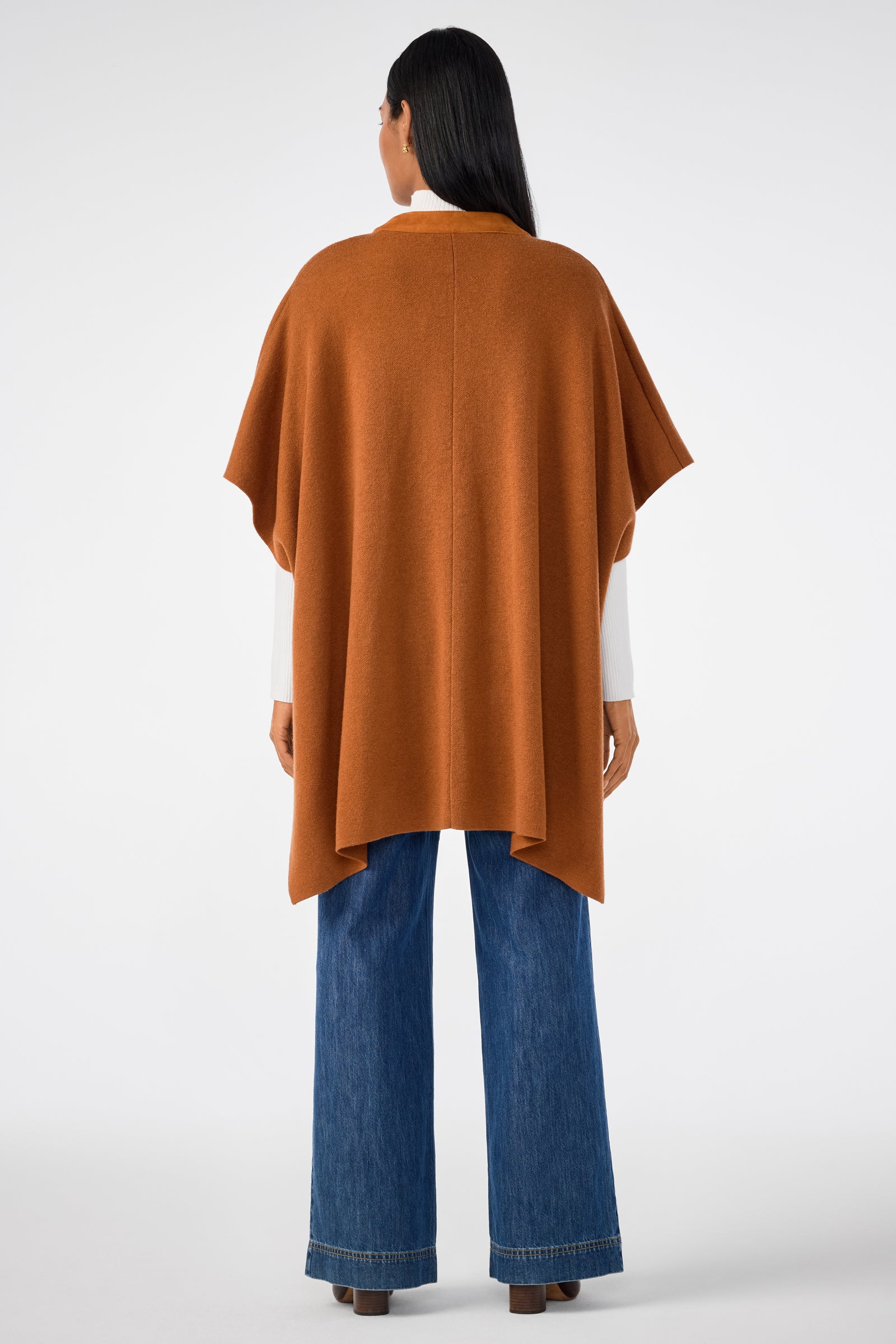 A woman faces away, wearing the Sapphire Poncho in rust over a white top and wide-leg blue jeans. Her straight dark hair falls past her shoulders against a plain white background.