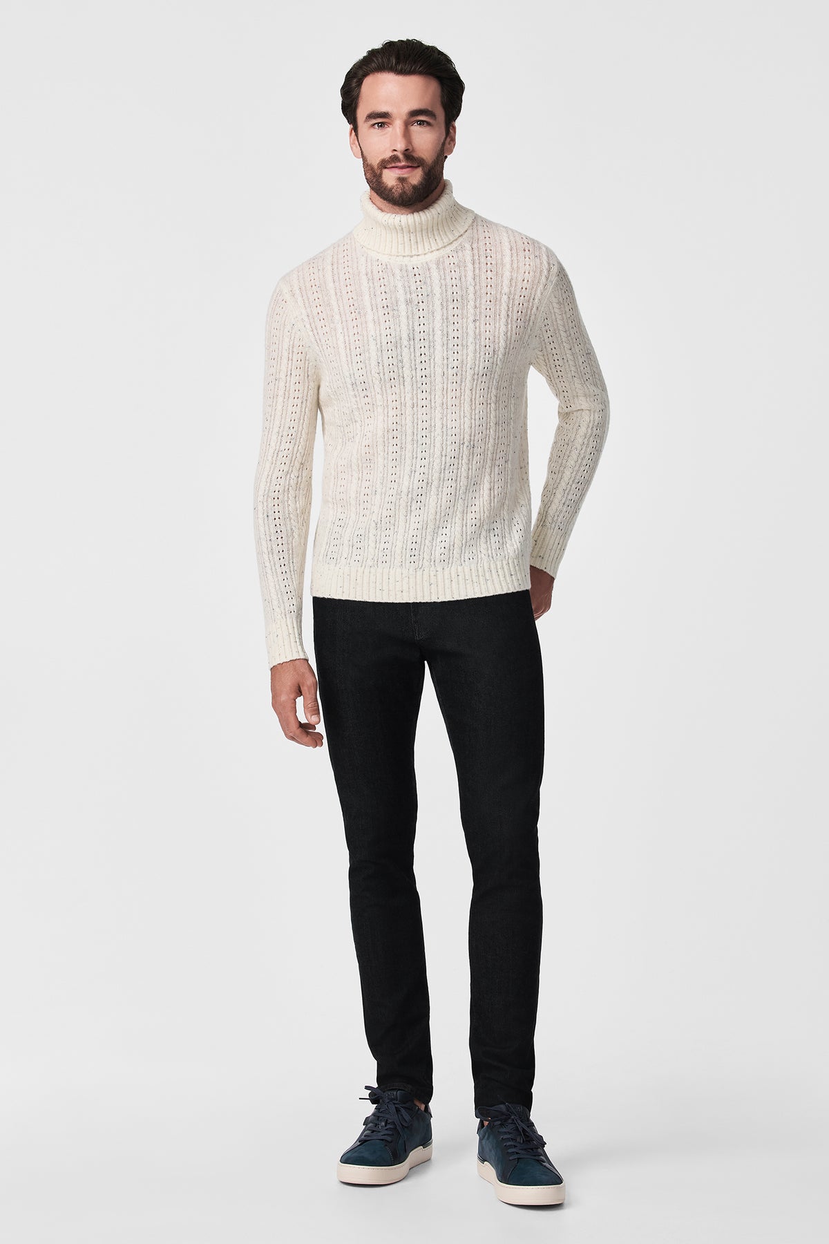 A man stands against a white background wearing the cream Wesley T-Neck sweater, black slim-fit pants, and navy blue sneakers.