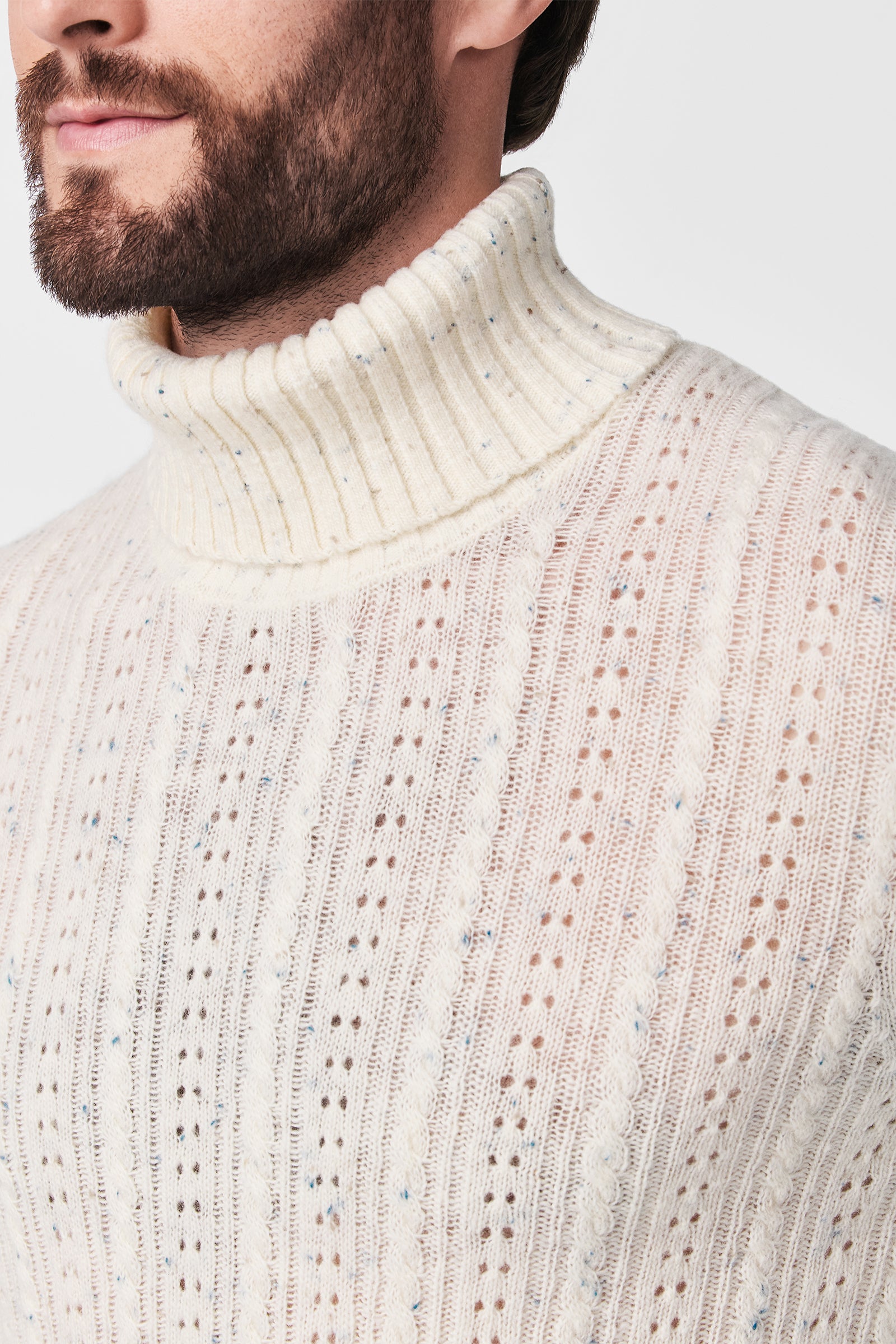 A close-up of a man wearing the Wesley T-Neck, a cream-colored, ribbed turtleneck sweater featuring a cable-knit and perforated texture. Only the lower half of his face, neck, and upper torso are shown.