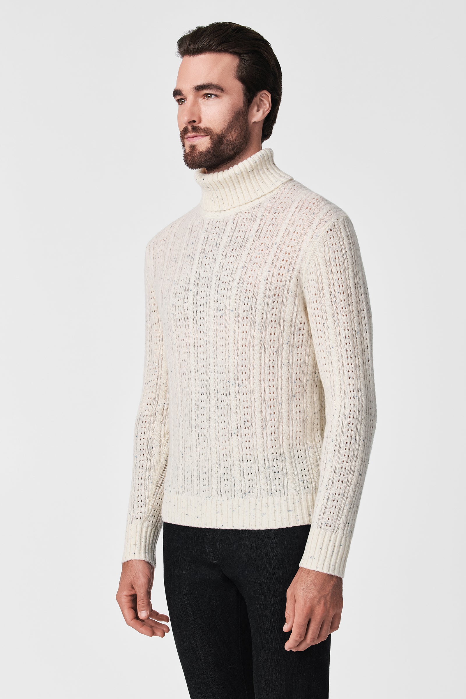 A man with a beard and dark hair models the Wesley T-Neck, a white textured turtleneck sweater, paired with black pants against a plain white background.