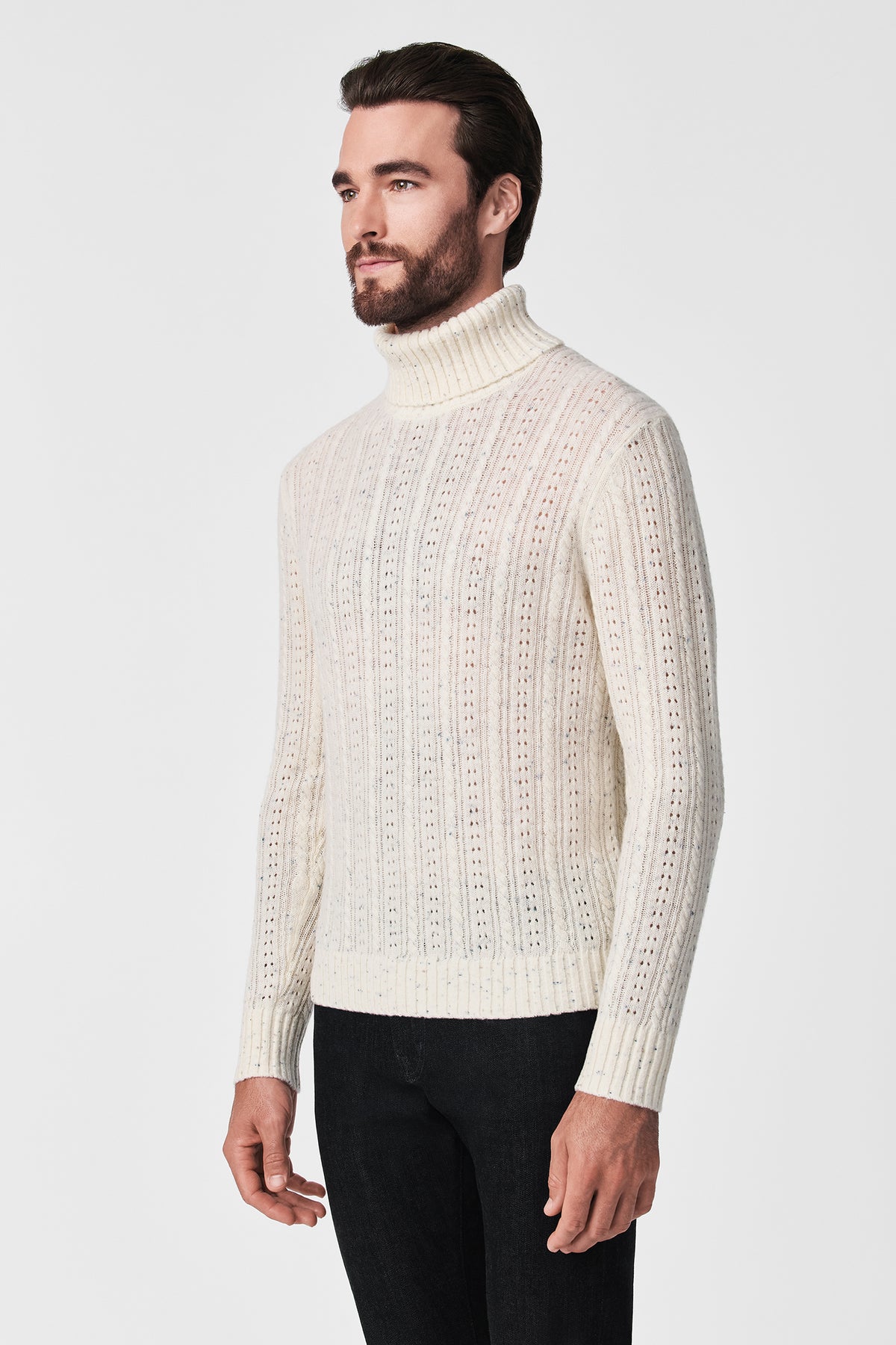 A man with a beard and dark hair models the Wesley T-Neck, a white textured turtleneck sweater, paired with black pants against a plain white background.