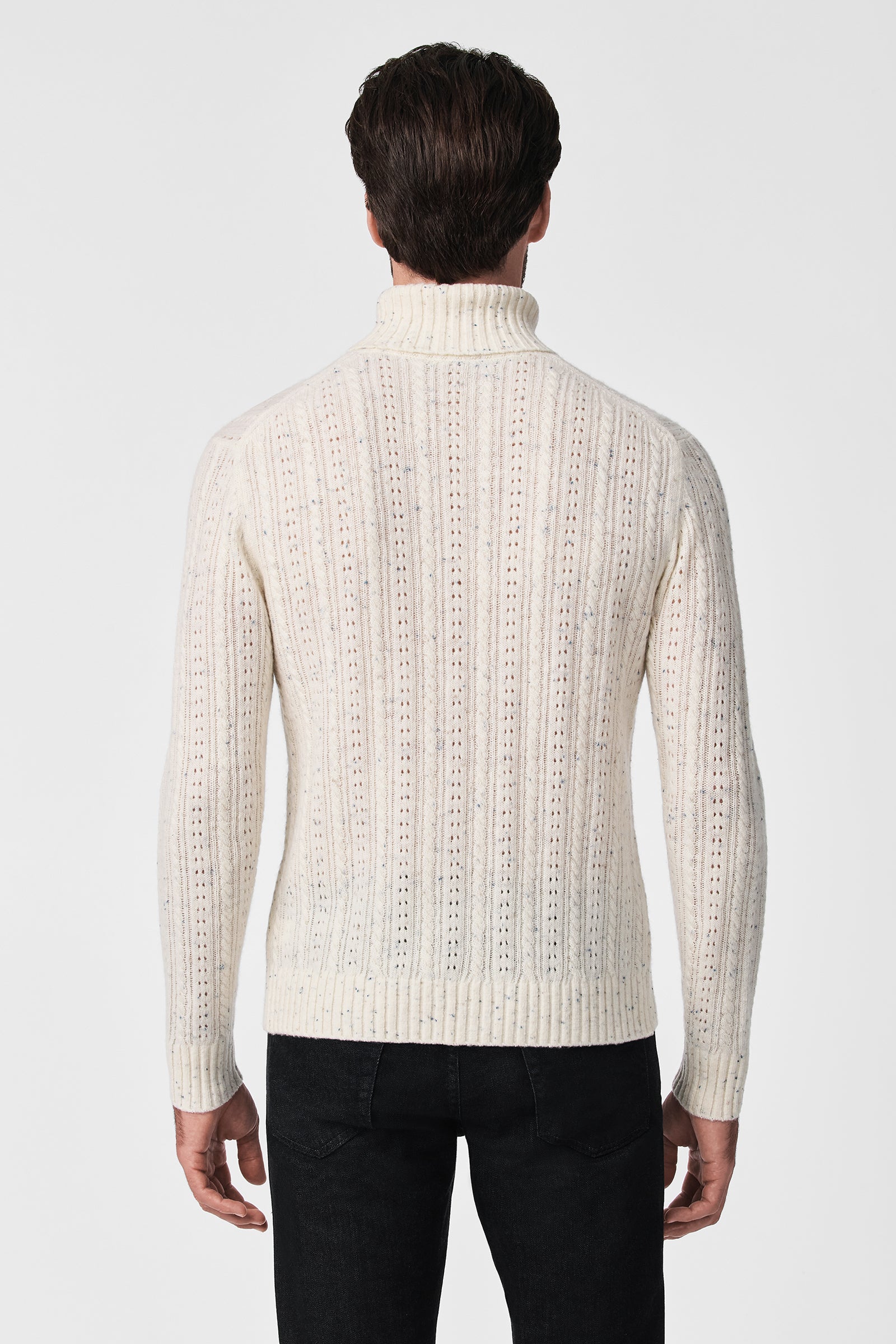 A man faces away, wearing the Wesley T-Neck—a cream-colored, textured knit turtleneck sweater with vertical ribbing and subtle patterns—paired with black pants. The background is a plain white.