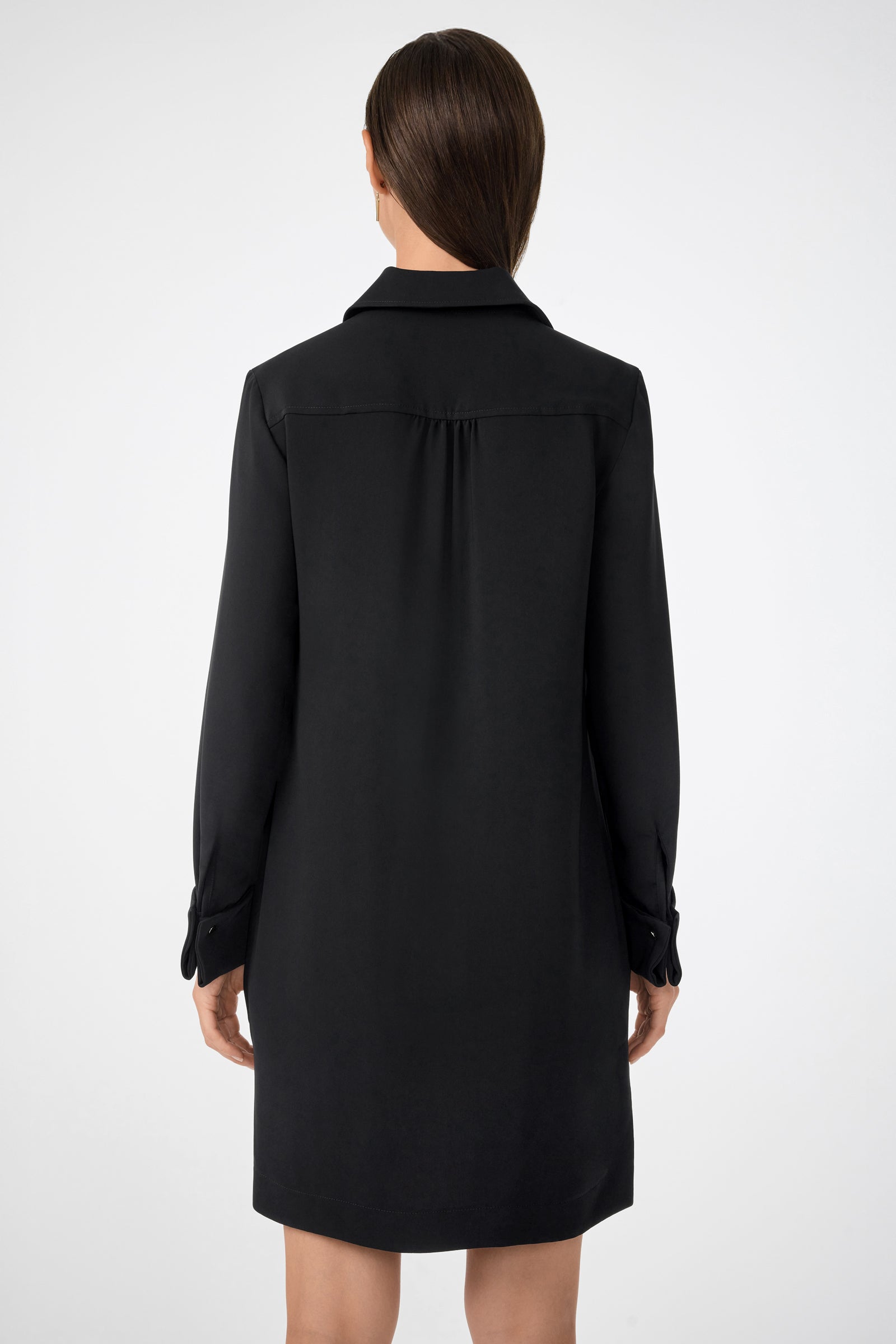 A woman with straight brown hair, seen from behind, wears the Darya Long-Sleeve Dress—black, knee-length—standing against a plain light background.