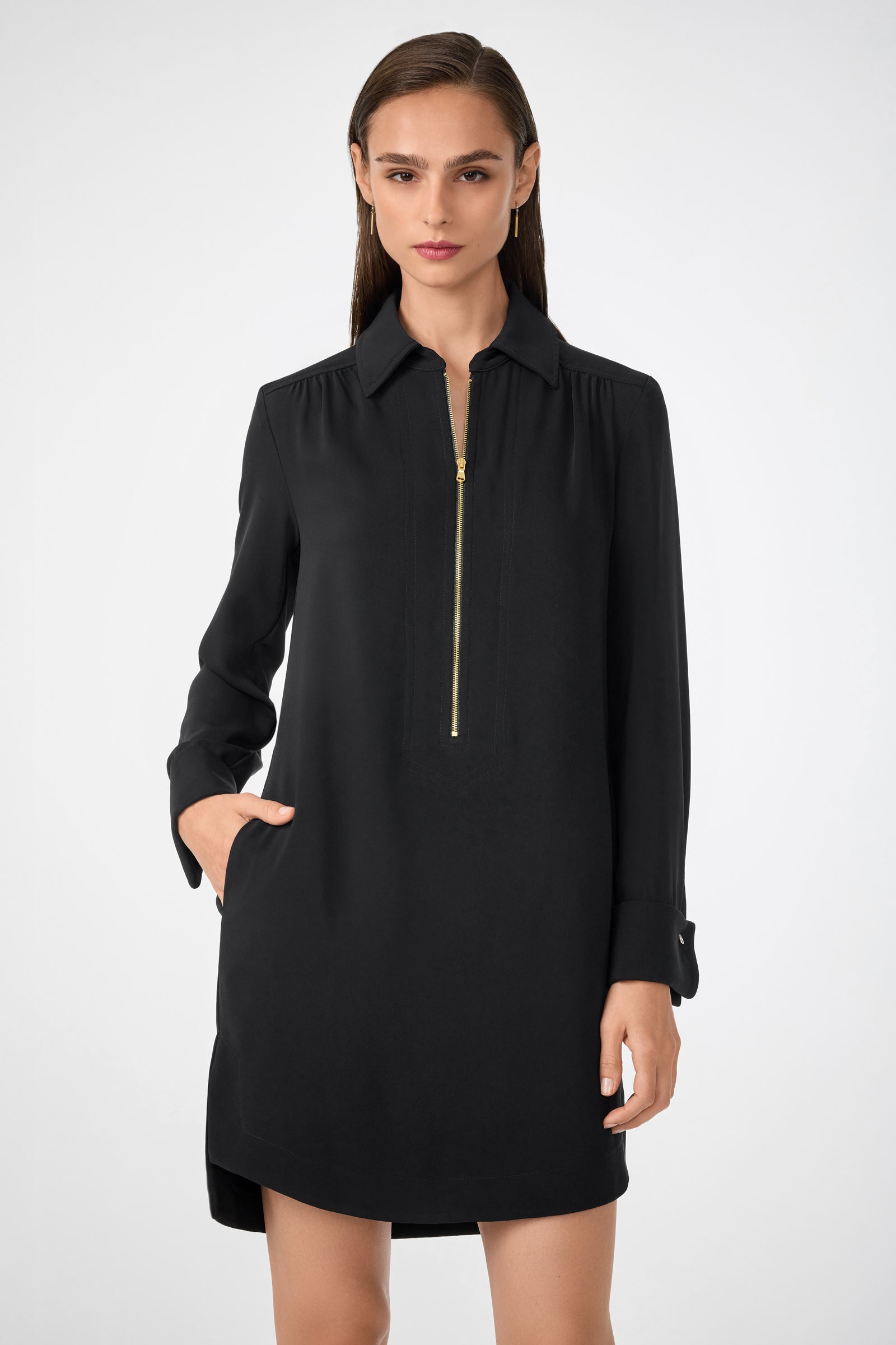 A woman wearing the Darya Long-Sleeve Dress—black, with a collar, gold front zipper, and side pockets—stands against a plain light background.