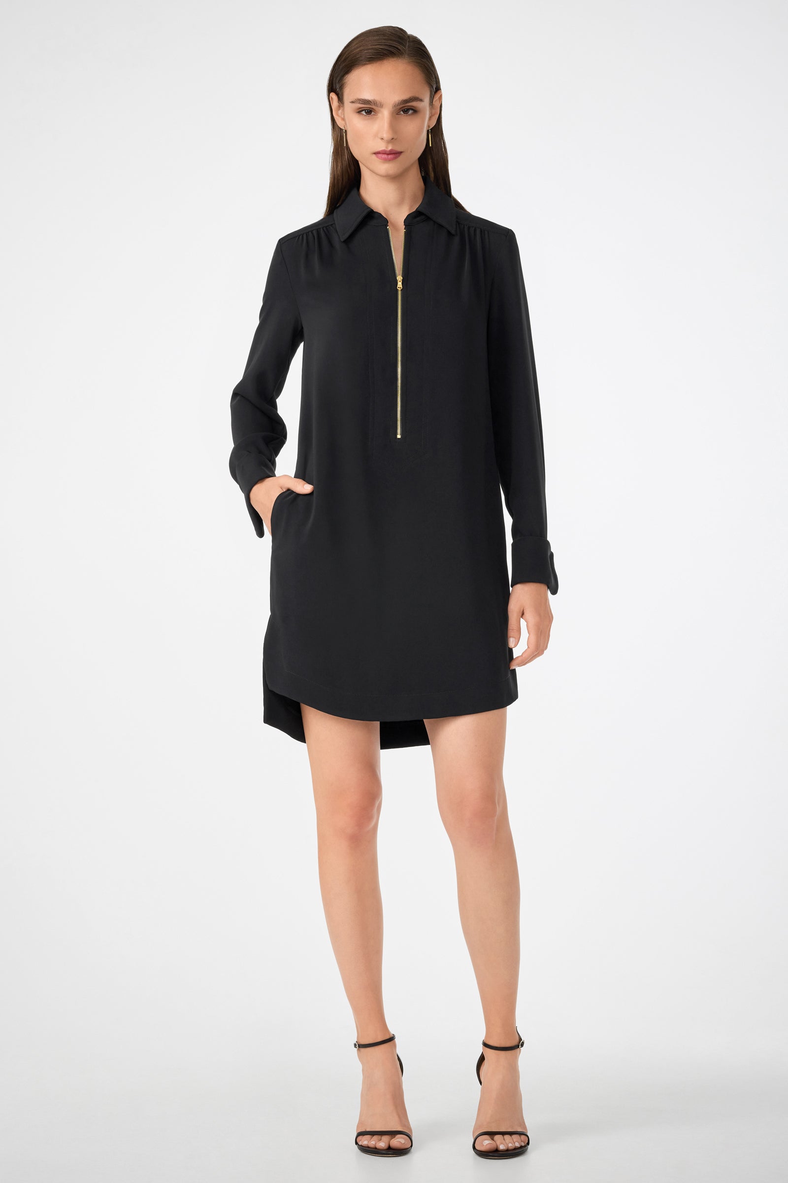 A woman with straight brown hair poses confidently in the Darya Long-Sleeve Dress in black, featuring a gold front zipper, side pockets, and black strappy heels against a plain background.