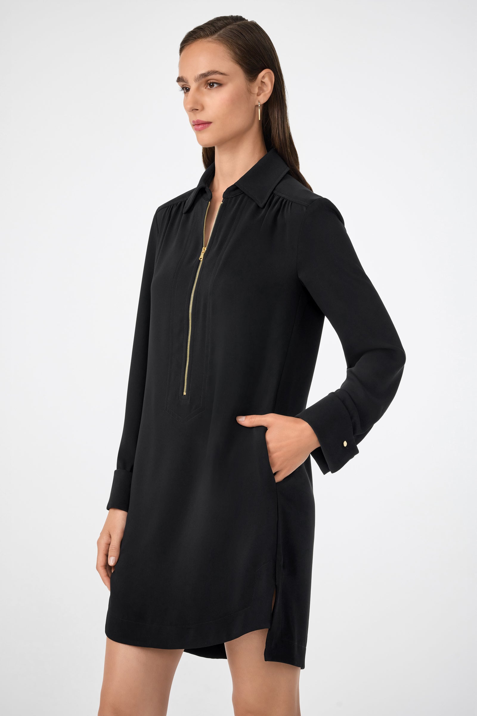 A woman with straight brown hair and a neutral expression stands against a plain background wearing the Darya Long-Sleeve Dress, featuring black fabric, a gold front zipper detail, and side pockets.