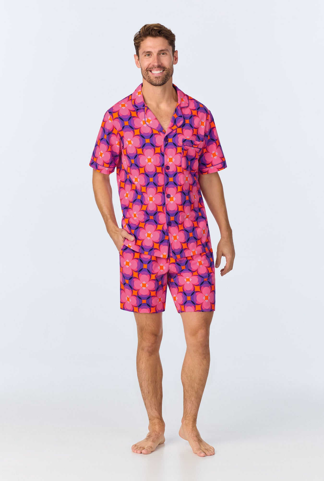 A man stands barefoot, smiling, wearing the Space Cat Mens Short Sleeve Boxer Short Jersey PJ Set in a vibrant multi-color design.