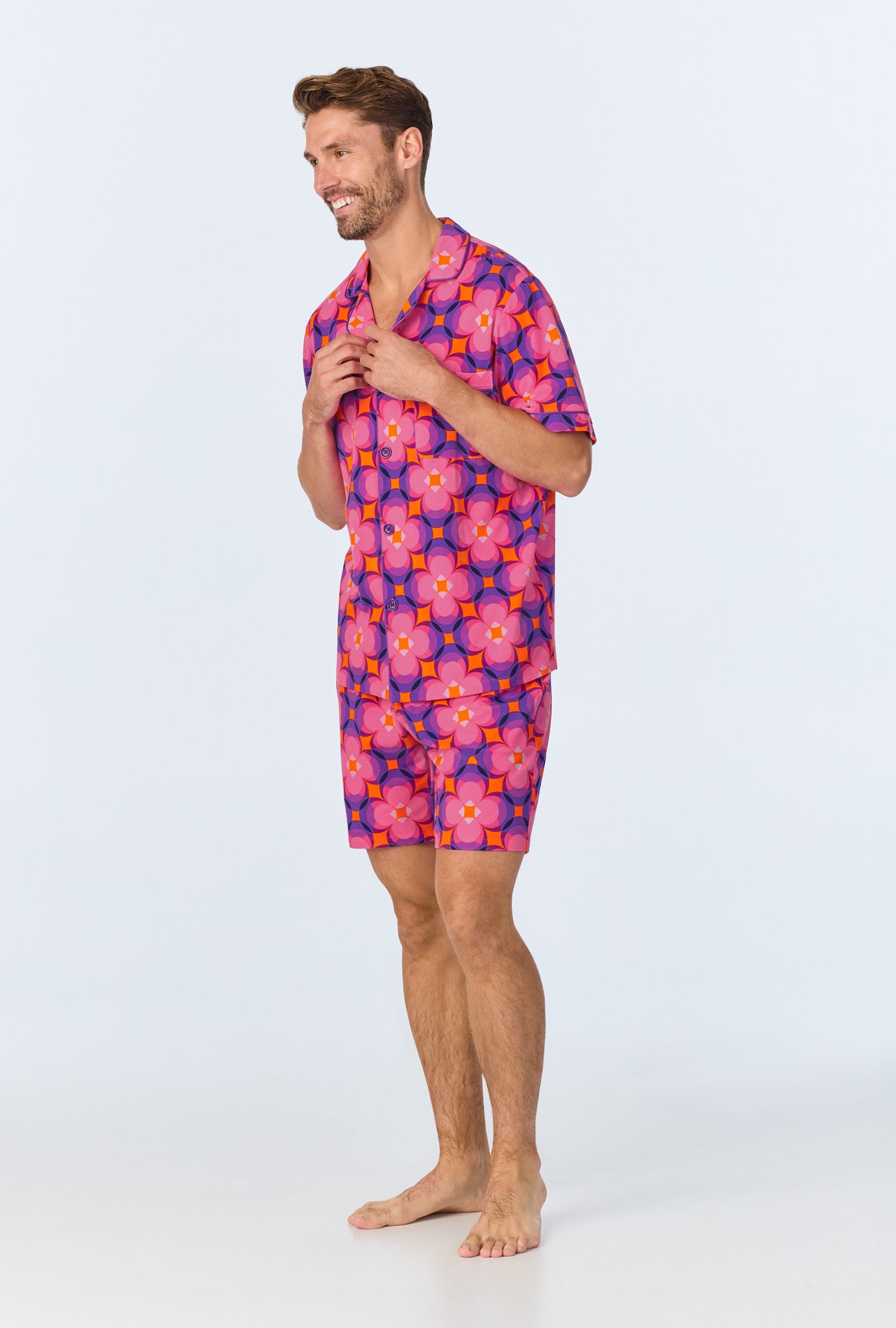 A smiling man stands barefoot in the Space Cat Mens Short Sleeve Boxer Short Jersey PJ Set, featuring a vibrant pink and purple floral print, made from soft organic cotton, against a plain light background.