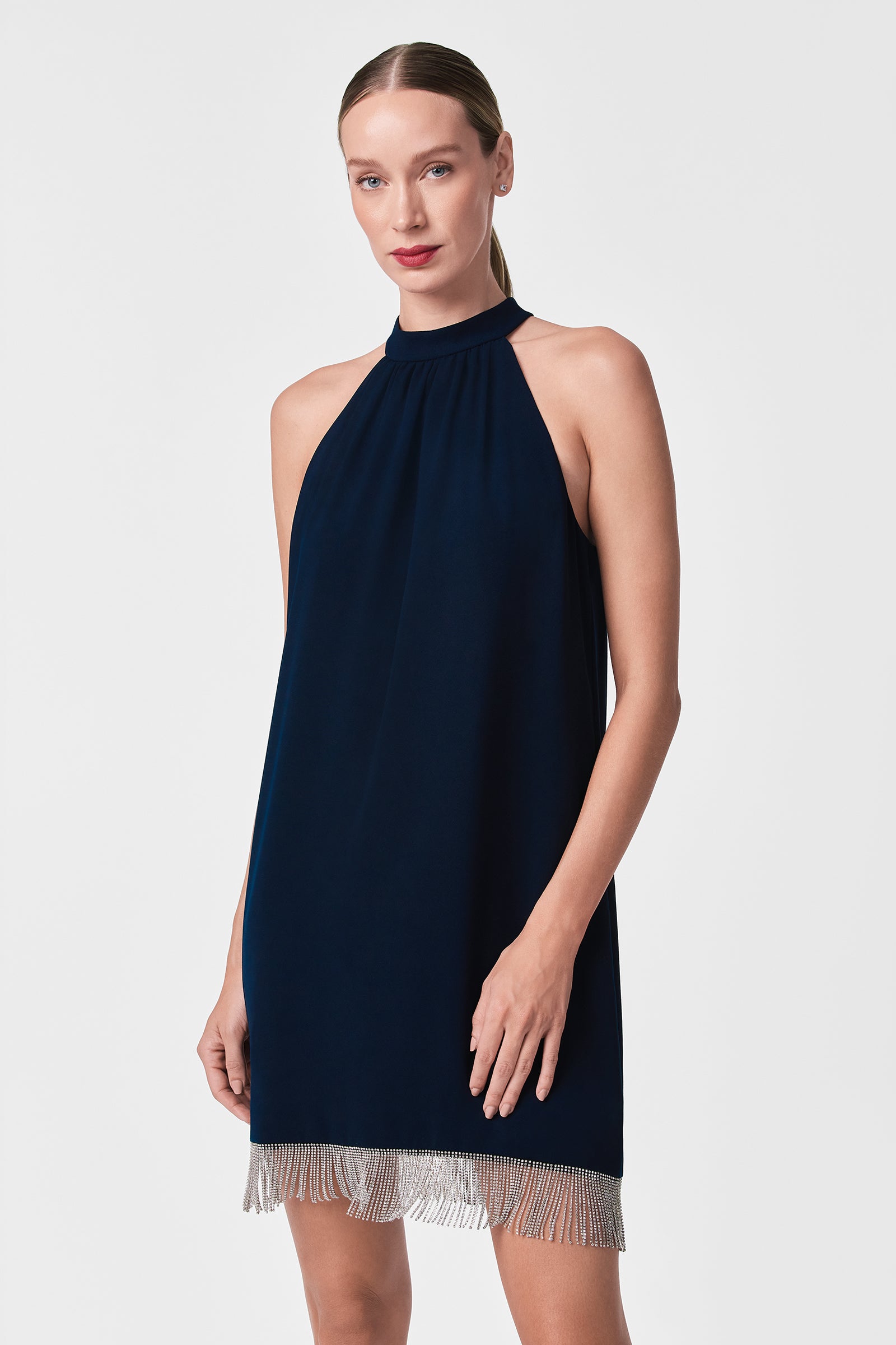 A woman stands against a plain white background, looking forward with her hair tied back, wearing the Top Shelf Dress—a sleeveless, dark blue halter-neck design featuring silver fringe at the hem.