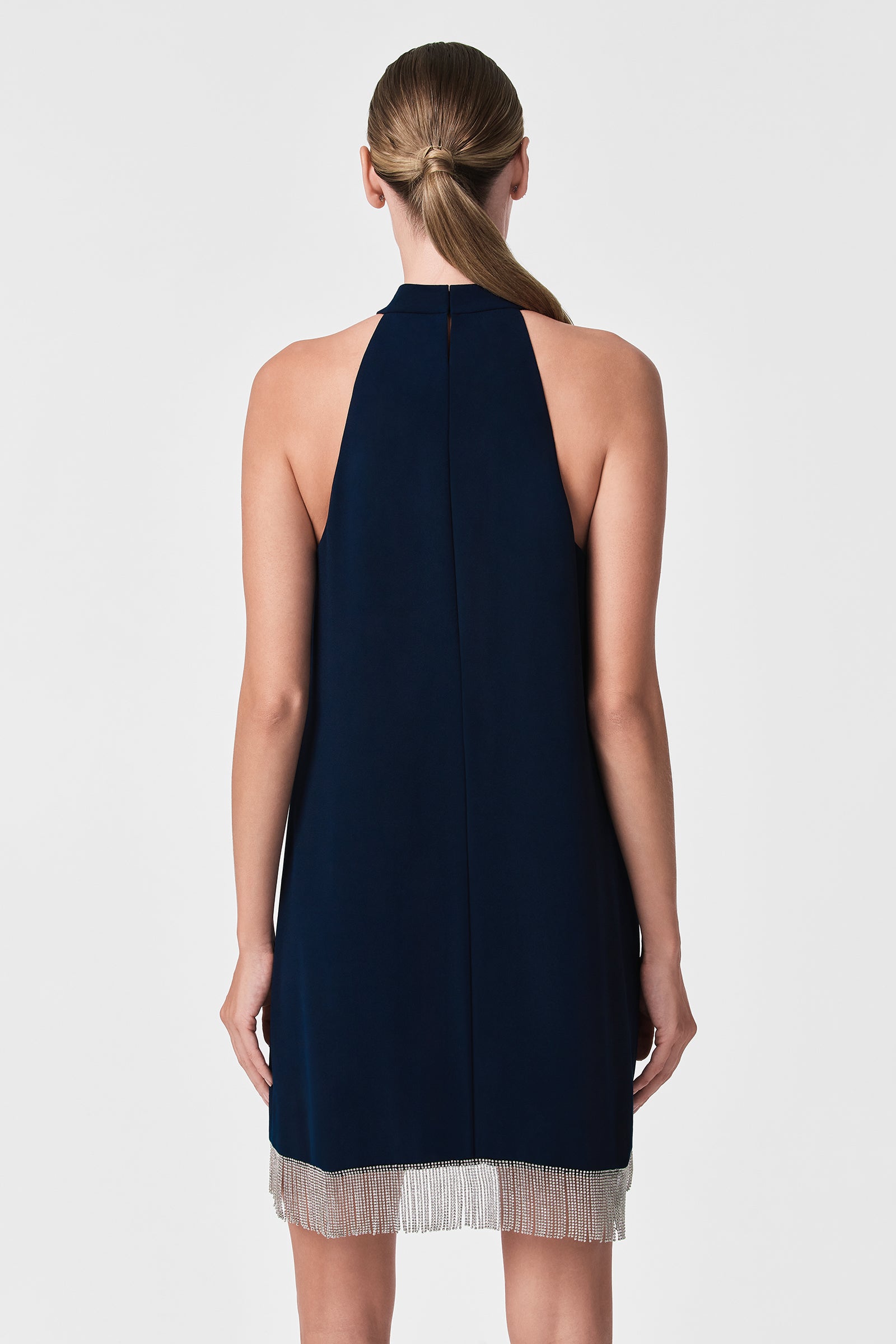 A woman with straight, light brown hair in a low ponytail wears the Top Shelf Dress—a sleeveless, navy blue dress with a high neckline and silver fringe trim—shown from the back against a plain white background.