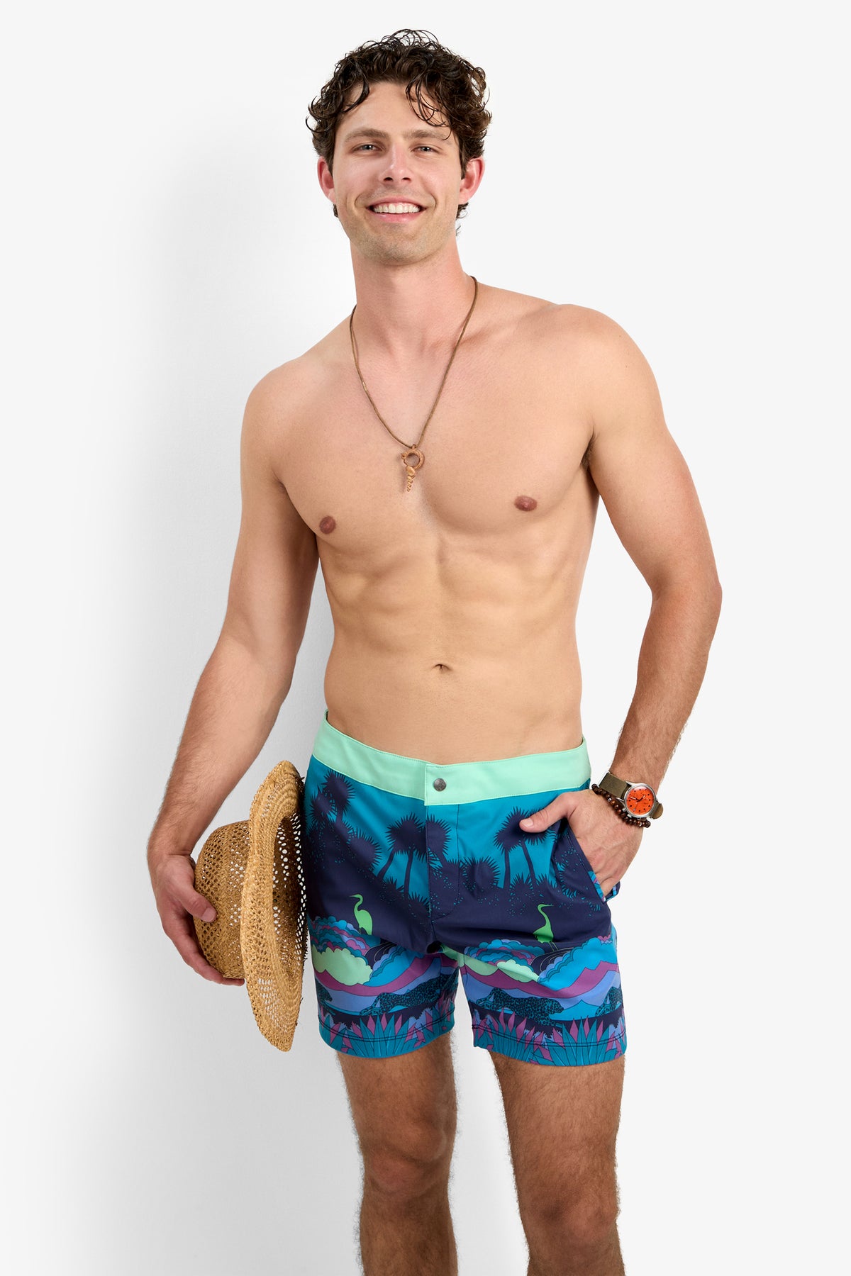 A smiling, shirtless man with curly hair wears the Honolulu Board Short - Jungle. He holds a straw hat, and accessorizes with a necklace and watch. The background is plain white.