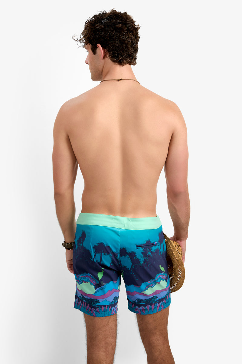 A man with curly hair stands shirtless, facing away, wearing the Honolulu Board Short - Jungle—turquoise and blue swim trunks with a tropical print—holding a straw hat in his right hand against a white background.