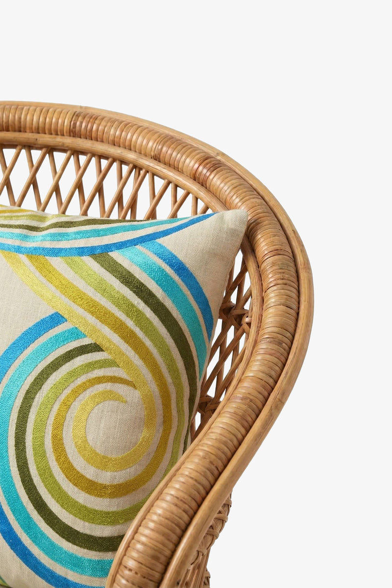A close-up of a rattan chair with a cream-colored 20″x20″ Calexico Embroidered Square Pillow, featuring blue, green, and yellow swirled patterns on a plain white background. The pillow cover is filled with plush down feathers for added comfort and support.
