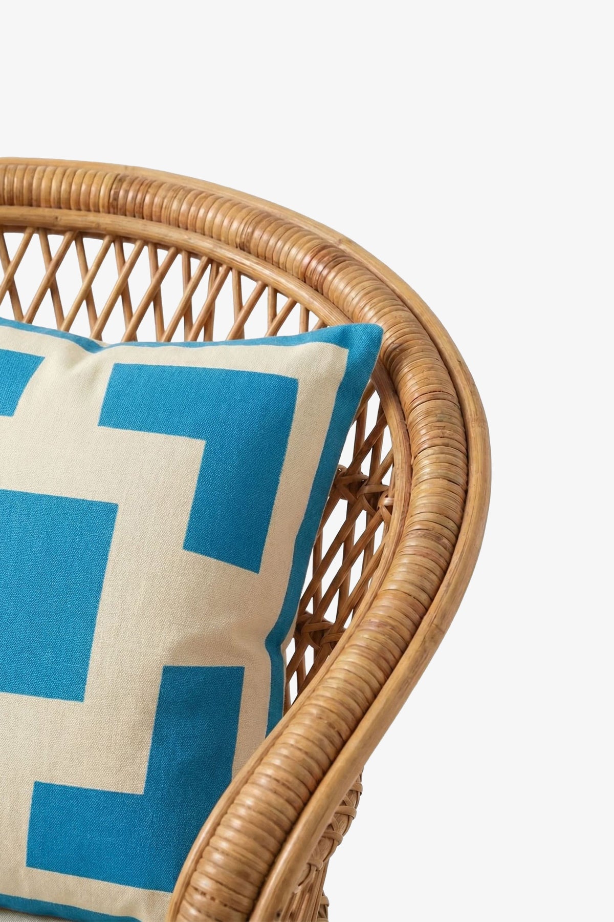 A close-up of a rattan chair with the Palm Springs Block Embroidered Square Pillow, featuring a 20″x20″ beige and blue geometric cover and a plush down feather insert, resting against its woven backrest on a plain white background.