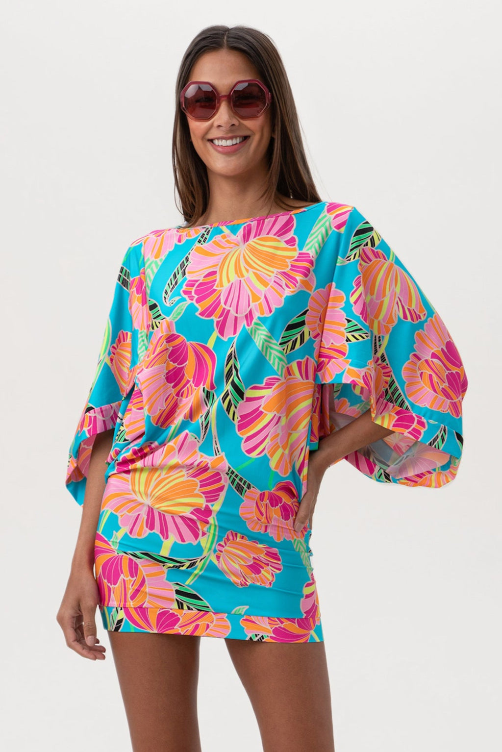 A woman in the Women's Poppy Boatneck Tunic Swim Cover-Up Dress, featuring large sunglasses and a vivid floral tropical print in pink, orange, and yellow on blue, smiles confidently against a plain white backdrop.