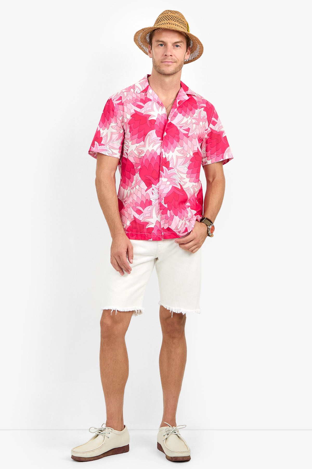 A man wearing the Felix Camp Shirt, white frayed shorts, light-colored shoes, and a straw hat stands smiling against a plain white background.