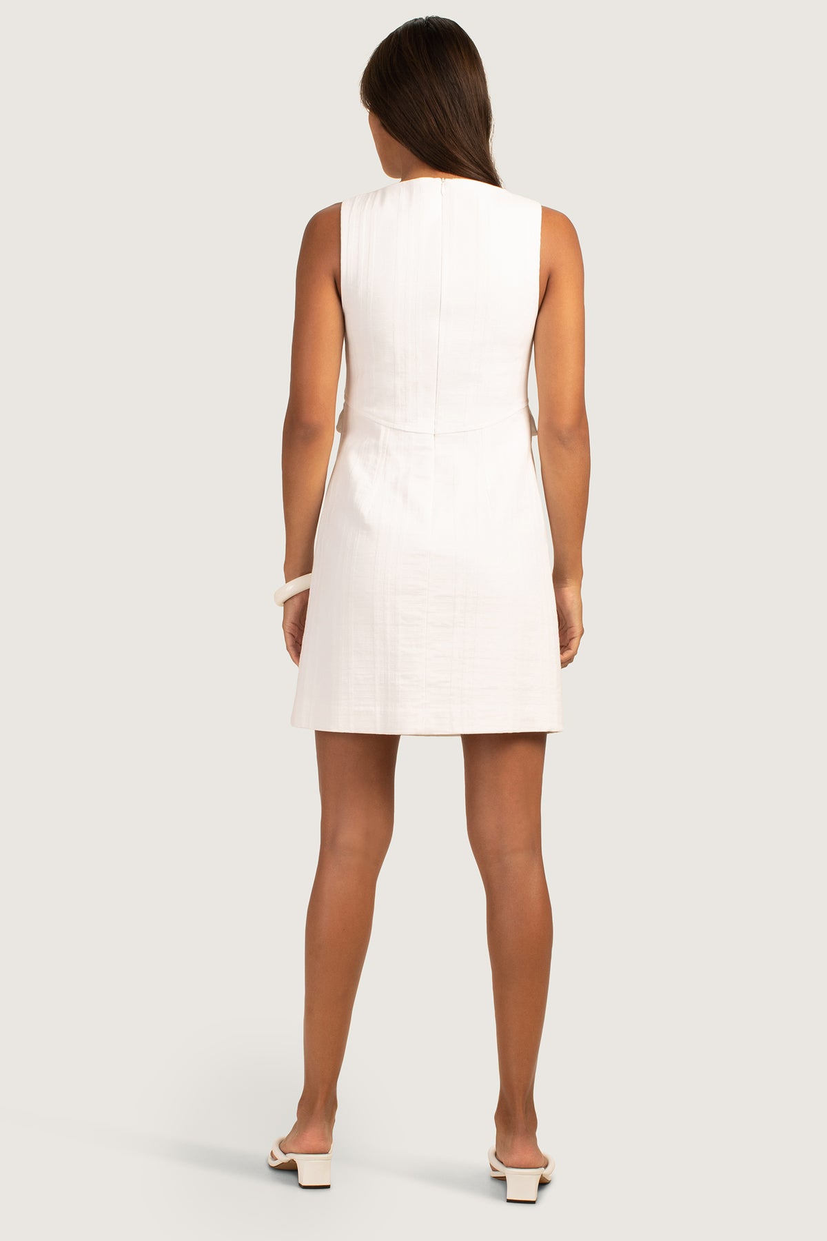 A woman with long brown hair, seen from behind, wears the sleeveless, knee-length white Atrina Dress with white heeled sandals against a plain light background.
