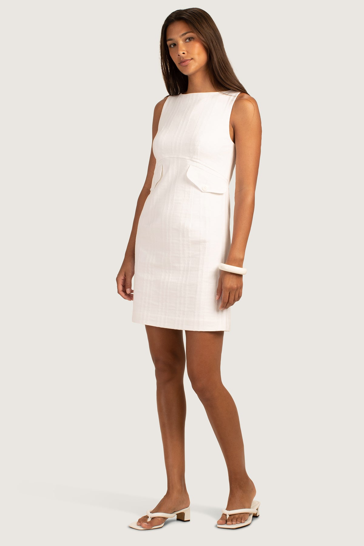 A woman poses against a plain light background, wearing the Atrina Dress—a sleeveless, knee-length boatneck sheath with a subtle check jacquard pattern—paired with white low-heeled sandals and a white bangle.