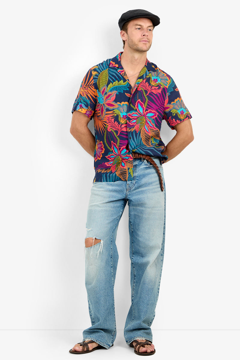 Man in a dark floral Felix Camp Shirt with a revere collar, light blue ripped jeans, brown sandals, and a black flat cap stands hands behind his back against a plain white background.