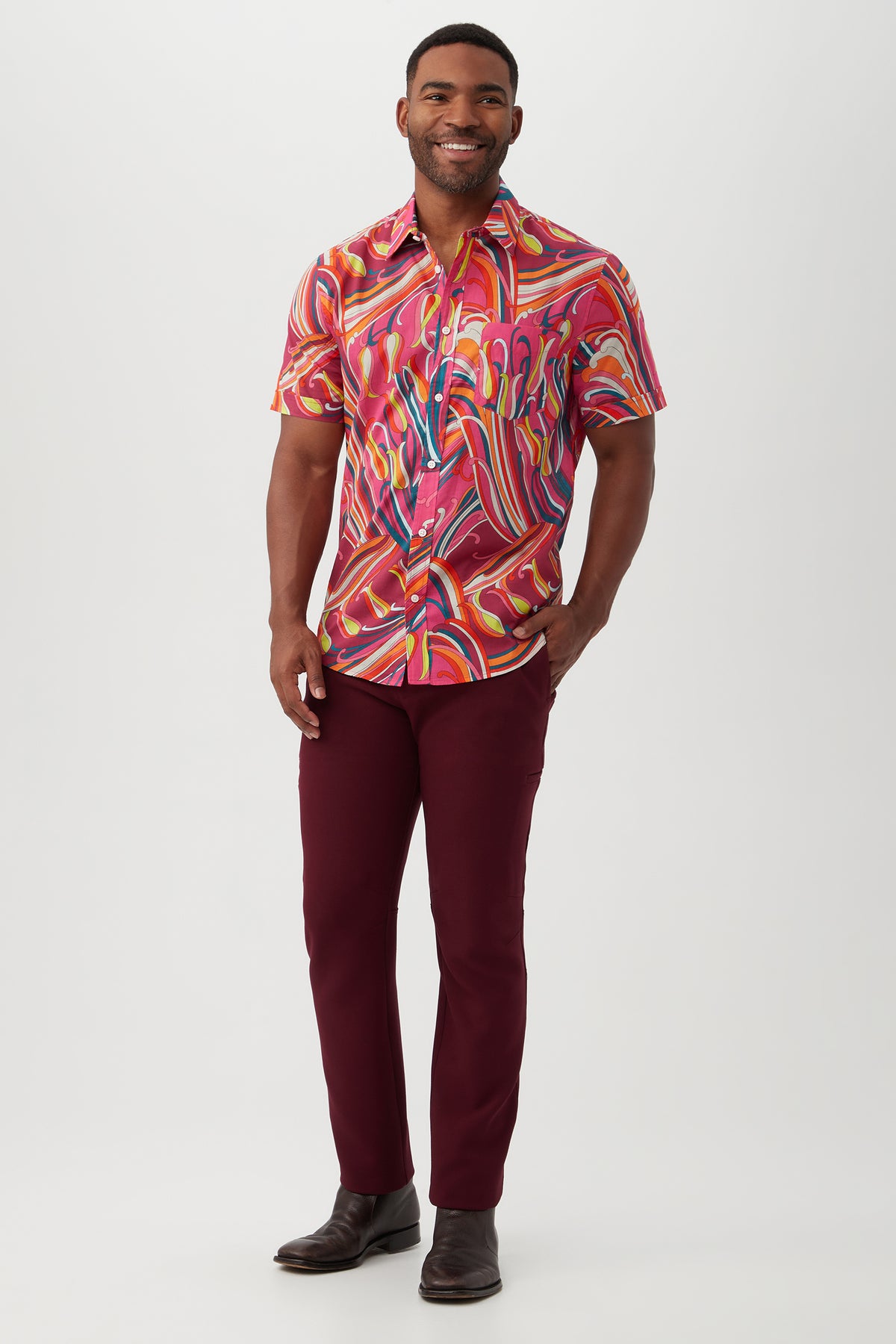 A man smiles while wearing the Slim Jim Shirt—a vibrant, swirling-patterned, short-sleeve button-up made from organic cotton—paired with dark red pants against a plain white background.