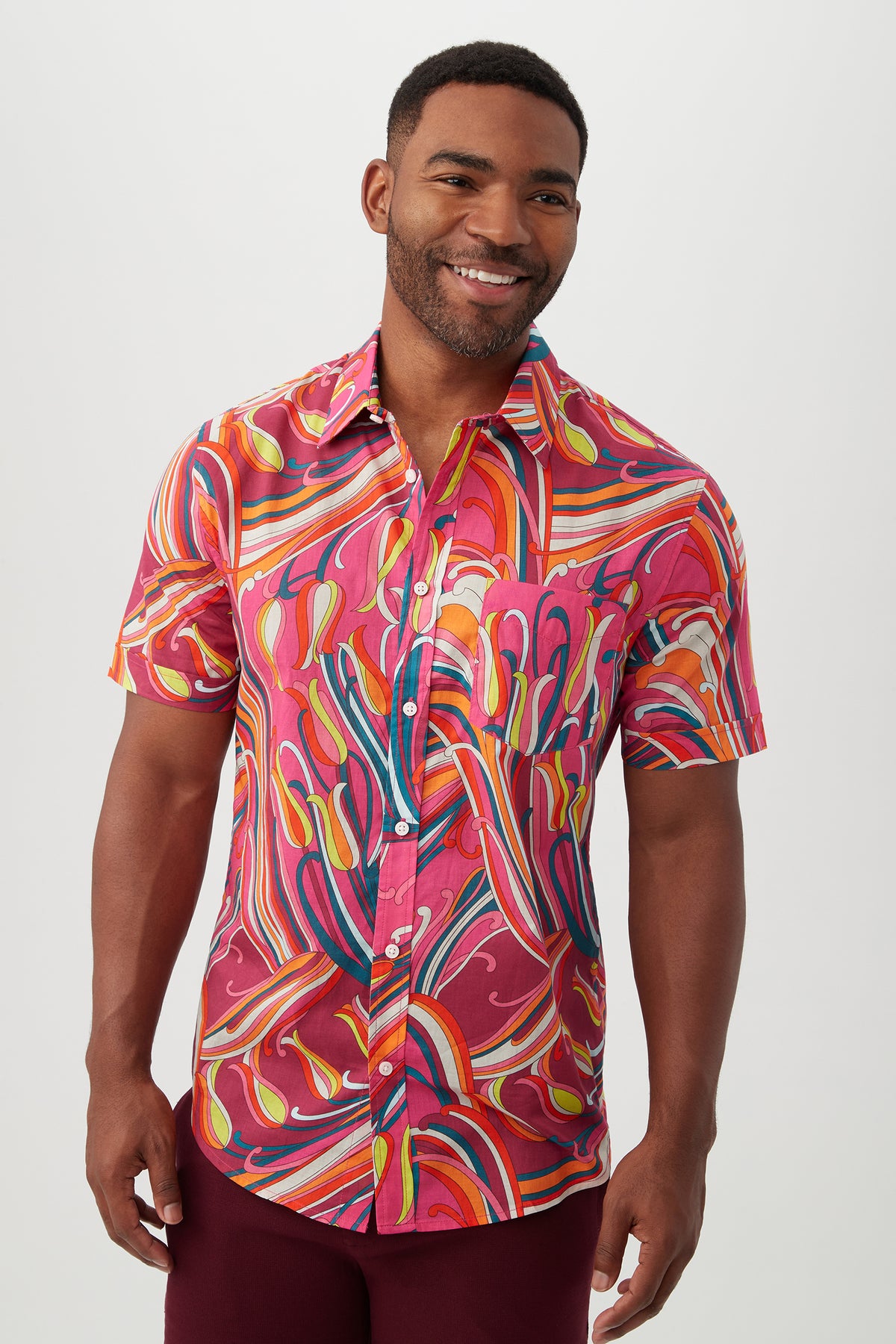 A man smiles in the Slim Jim Shirt, a slim fit, short-sleeve button-up in Radio City Rose Multi, featuring a bright swirling abstract pattern. He stands against a plain light background.
