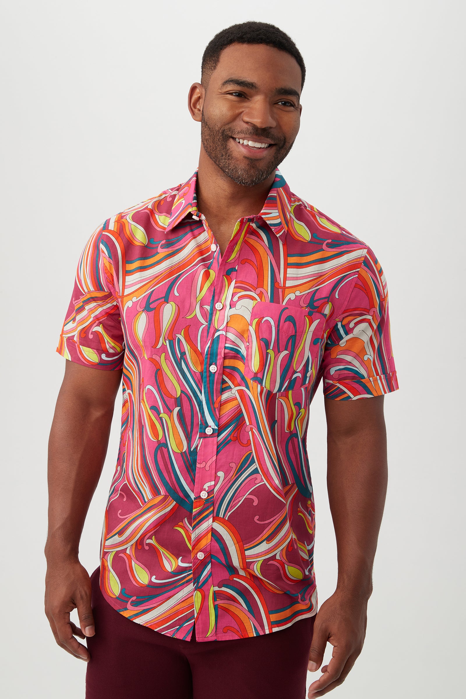 A man smiles in the Slim Jim Shirt, a slim fit, short-sleeve button-up in Radio City Rose Multi, featuring a bright swirling abstract pattern. He stands against a plain light background.