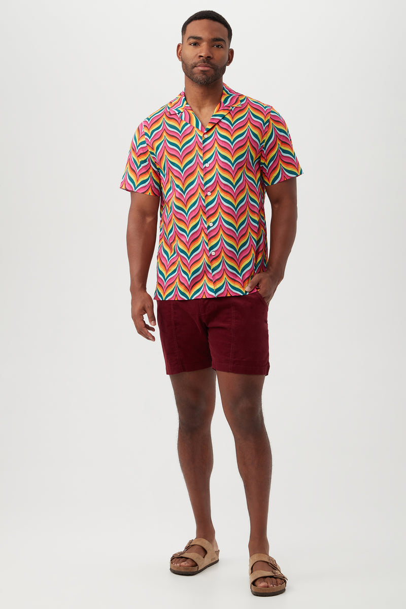 A man stands against a plain background wearing the Felix Shirt, which features a colorful zigzag pattern and revere collar, paired with maroon shorts and beige sandals, with one hand in his pocket.