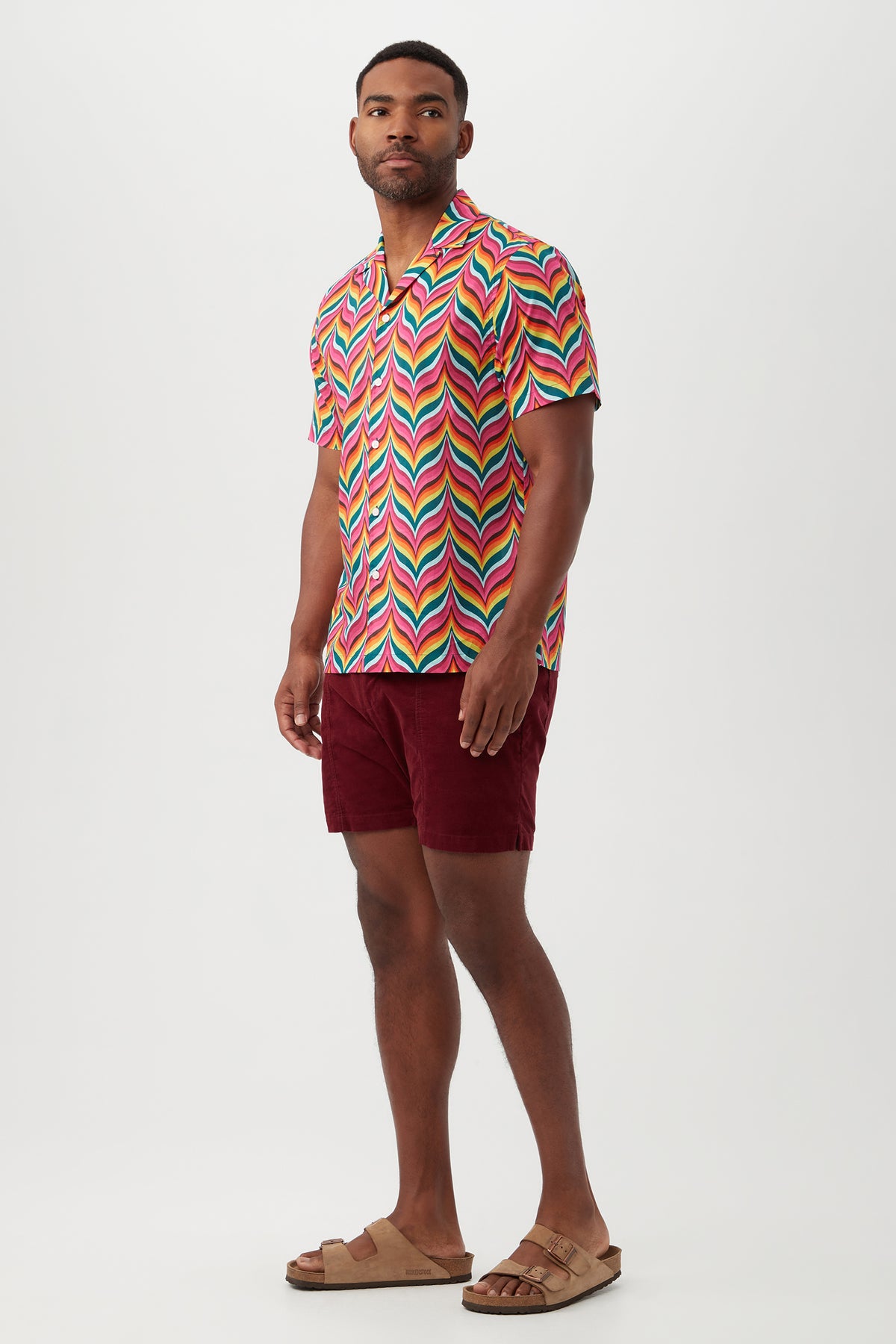 A man stands against a plain white background wearing the Felix Shirt, featuring a multicolored zigzag pattern and revere collar, paired with burgundy shorts and tan sandals.