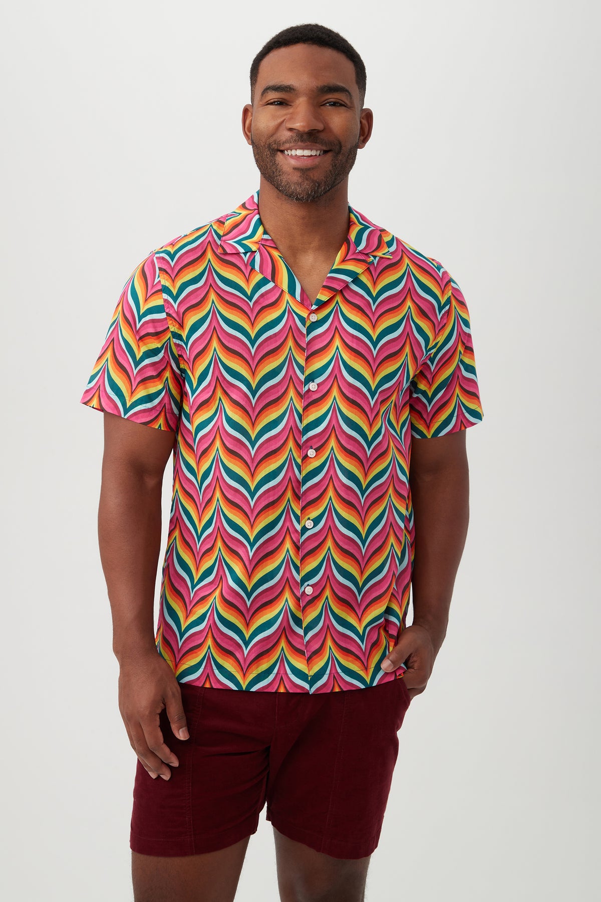 A man smiles while wearing the Felix Shirt in Multi, a brightly colored short-sleeve camp shirt with a bold rainbow zigzag pattern and revere collar, paired with dark red shorts, standing against a plain light background.
